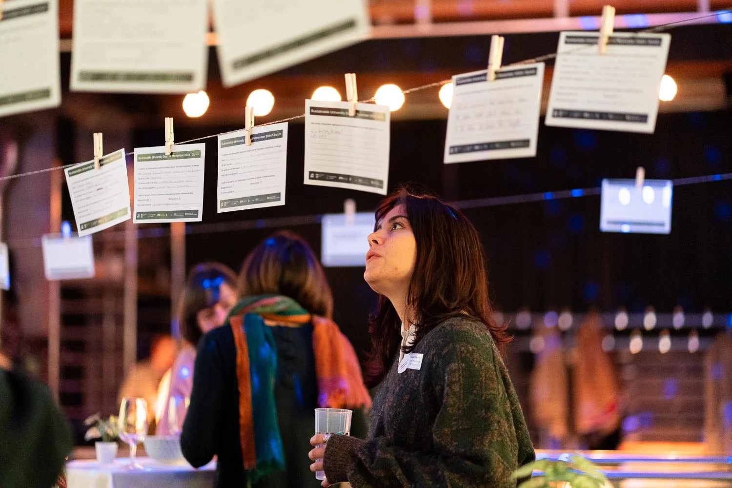A woman with dark hair and a brown sweater holding a glass, looking up at papers hanging on strings with clothespins at an indoor event.
