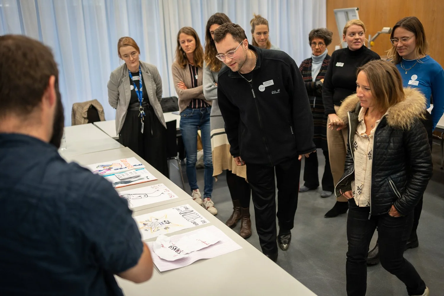 A group of people gathered around a table looking at drawings and papers with one person explaining or presenting.
