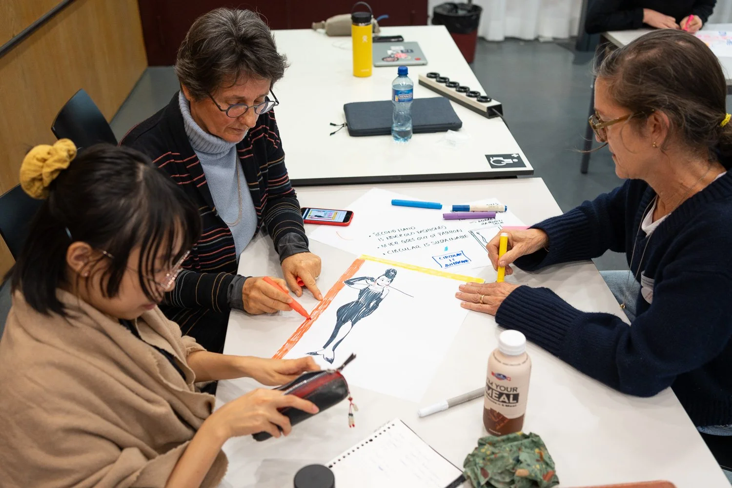 Three women are sitting at a table engaged in drawing and discussing a fashion illustration of a woman in a dress, with colored markers, paper, and sketch notes on the table.
