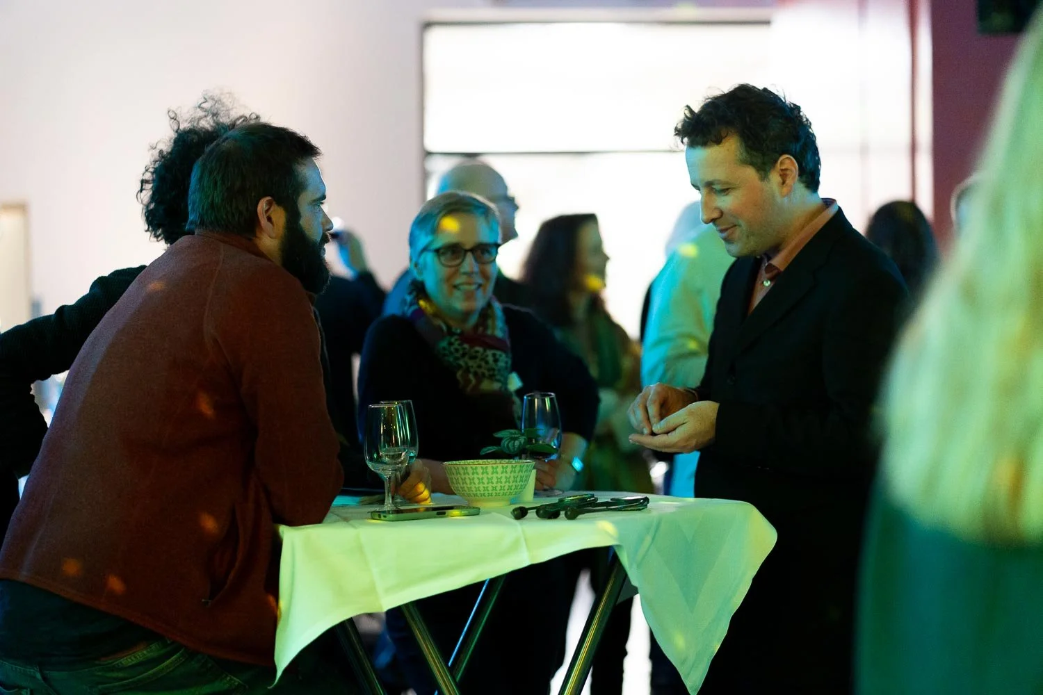 People having a conversation at a social event, standing around a cocktail table with glasses and a bowl.