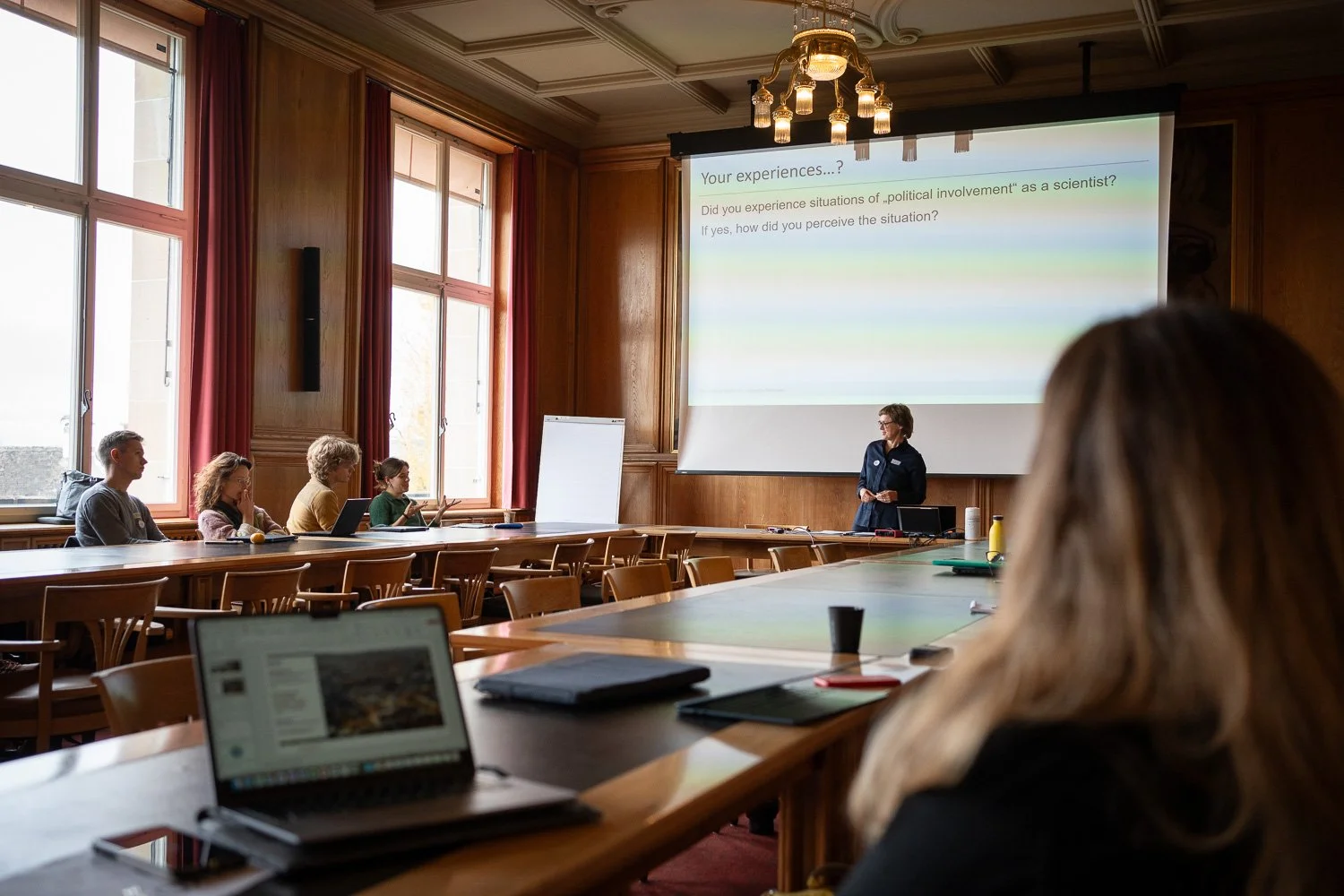 A woman giving a presentation in a large conference room with wooden walls and large windows, with a slide projected on a screen behind her that asks about experiences with political involvement as a scientist.