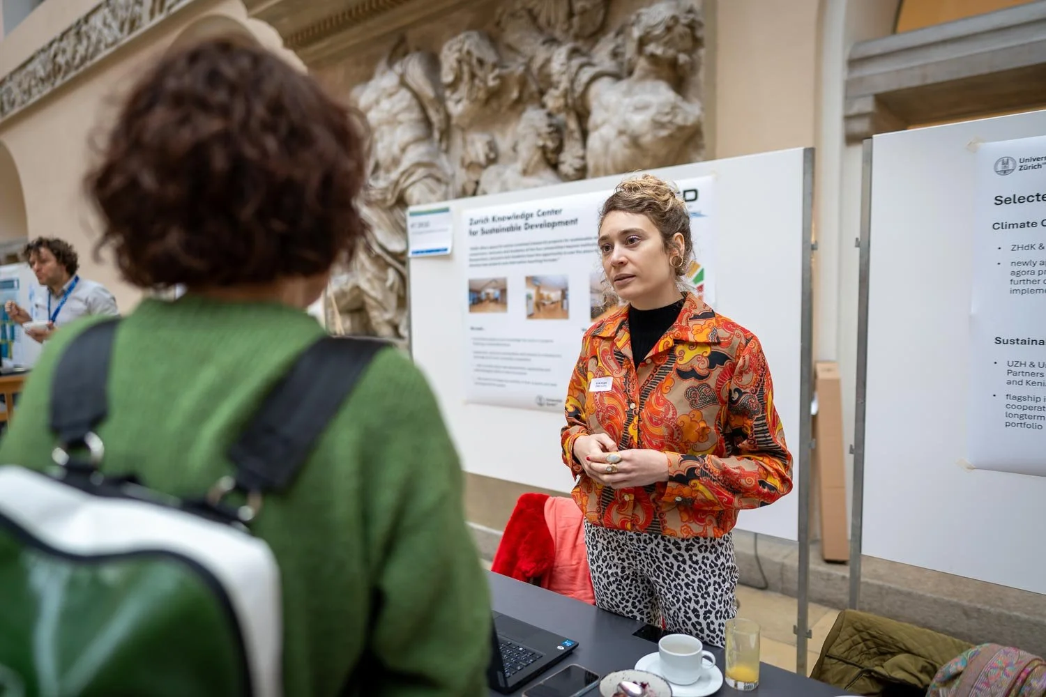 A woman with curly hair and a colorful orange and black patterned jacket is speaking to a woman with short brown hair, wearing a green sweater, at an indoor exhibition. The woman in the jacket is gesturing with her hands while the woman in green list