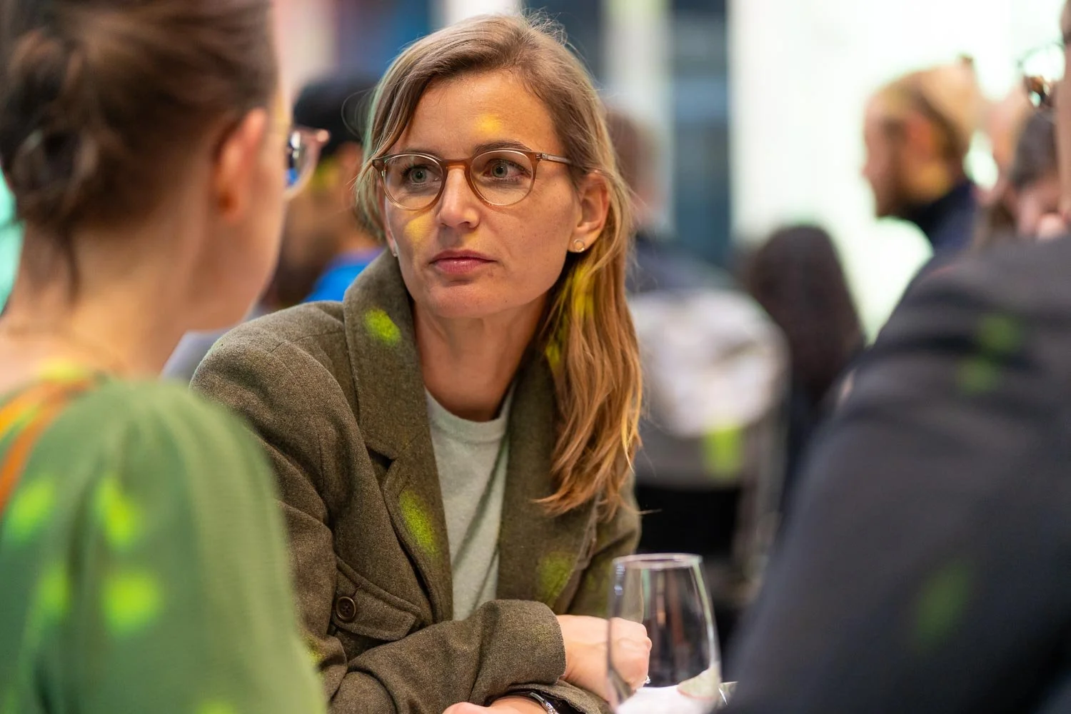 Women engaged in conversation at a social event, with a blurred background of other people, and a glass of wine on the table.