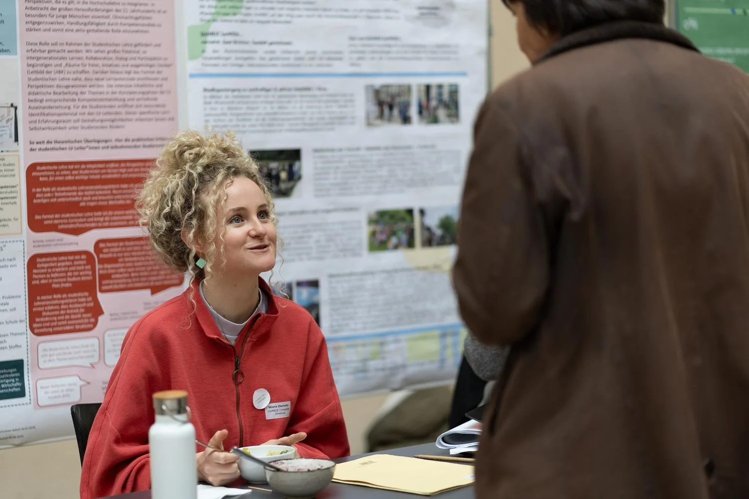 A young woman with curly blonde hair wearing a red jacket and a badge, sitting at a table with a bowl of food and a water bottle, talking to a person in a brown jacket against a backdrop of informational posters.