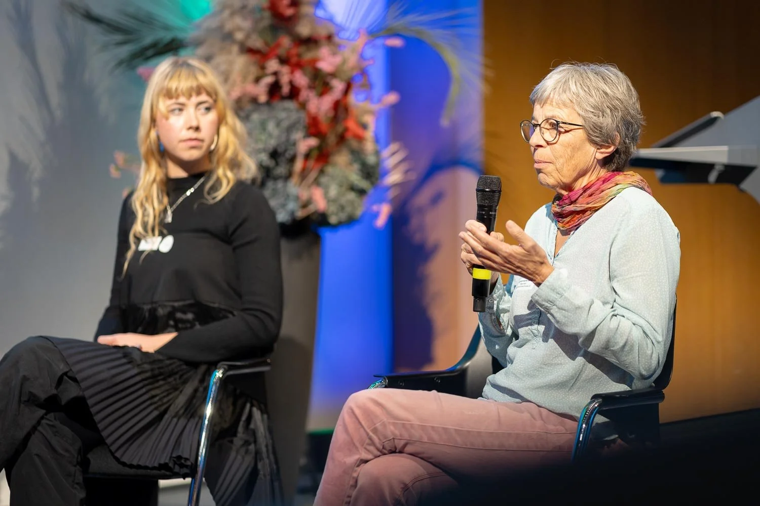 An older woman with glasses and gray hair speaking into a microphone, sitting next to a young woman with blonde hair listening attentively. They are on stage with a floral arrangement in the background.