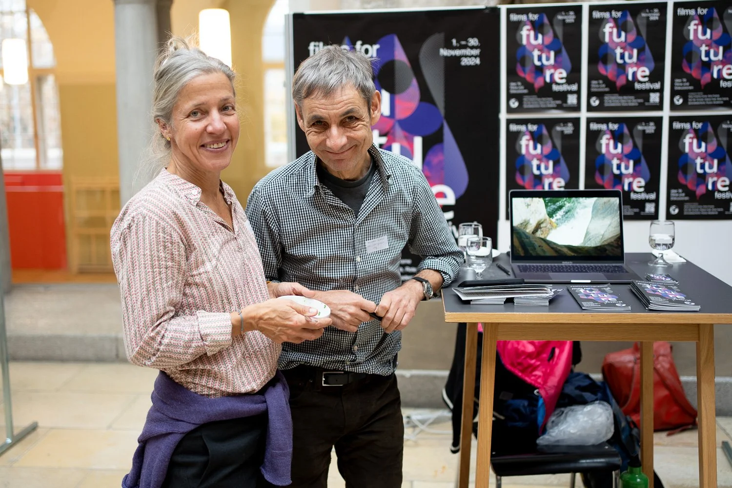 A woman and a man standing together at an event, smiling at the camera. Behind them is a display with multiple posters for a film festival called "Film for Future," featuring a colorful abstract design, a row of water glasses, a laptop, and informati