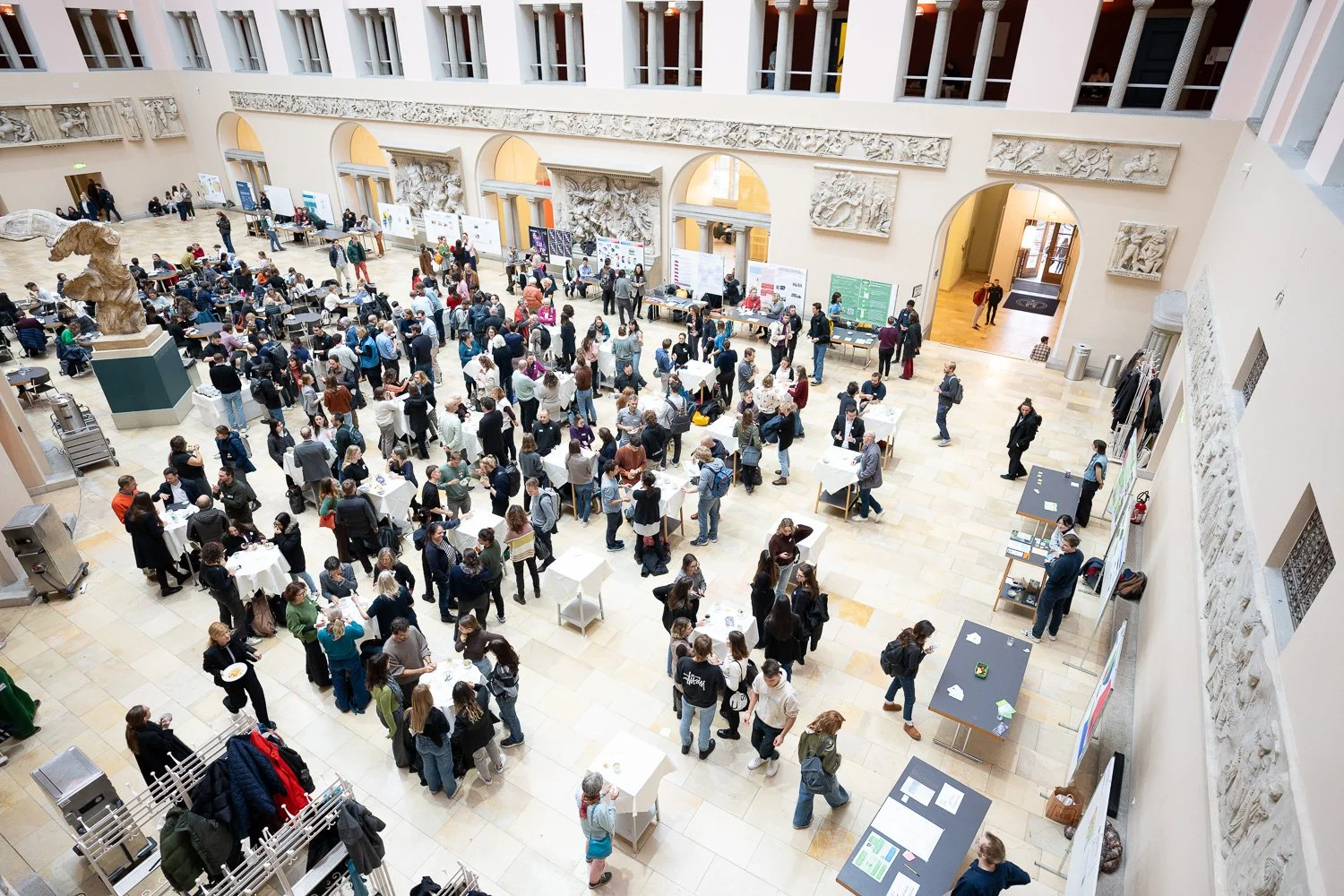 A large crowd of people attending an event in a spacious, historic-style building with high ceilings and decorative reliefs on the walls, many of them standing near tables or walking around.