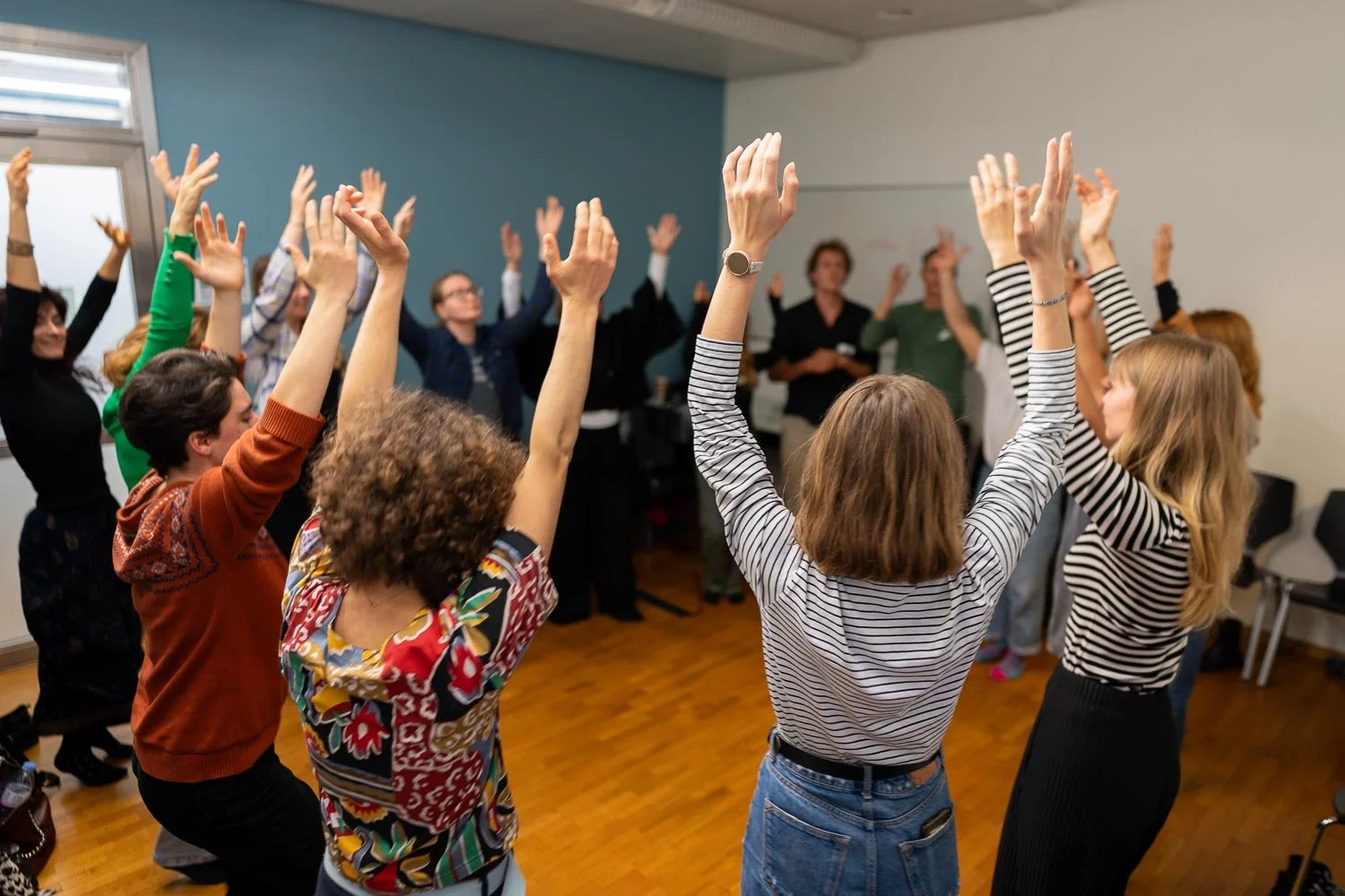 Group of women participating in a dance or exercise class indoors, raising their hands in the air, on a wooden floor, with some chairs against the wall.