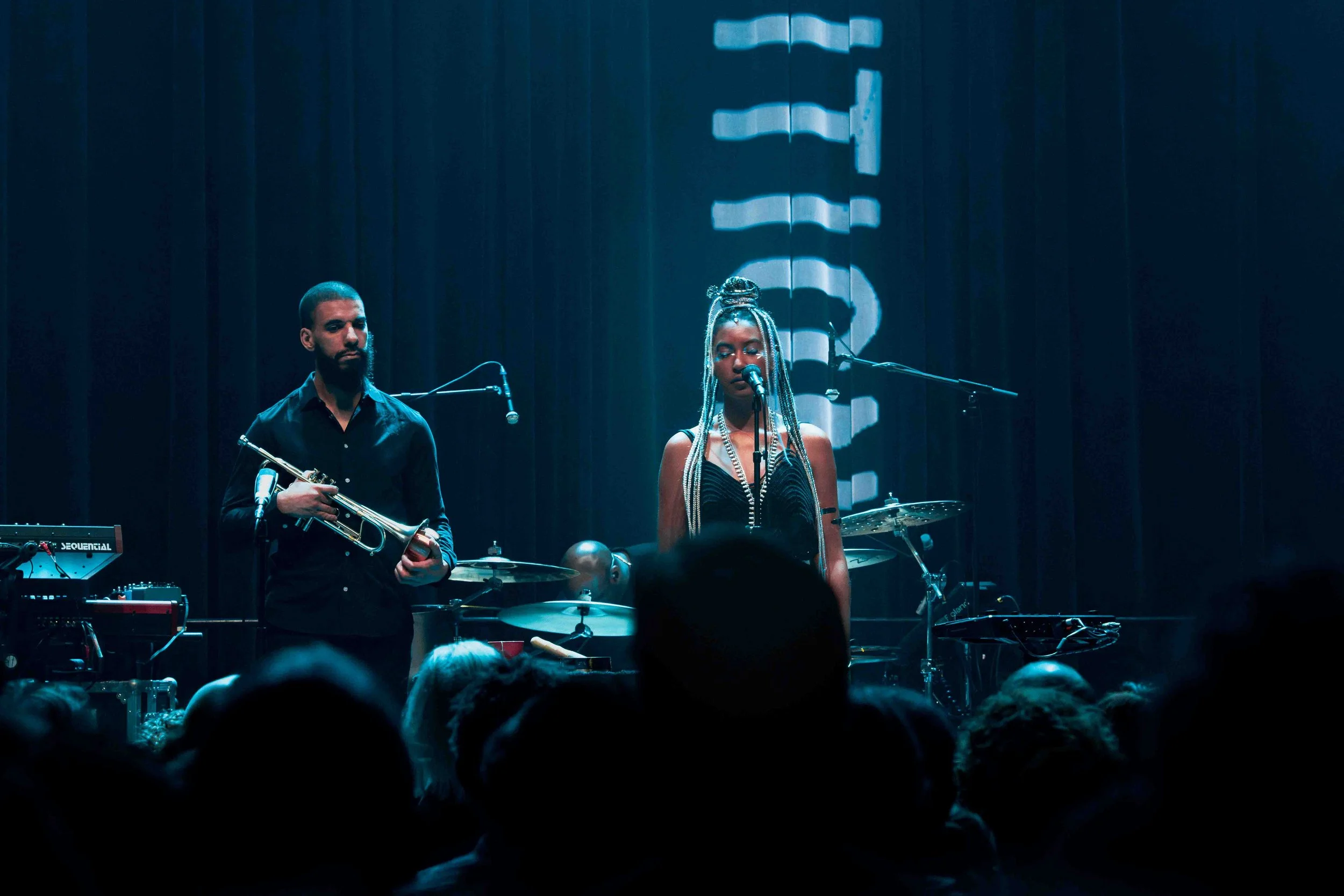 Muziekband op een podium tijdens een live optreden, bestaande uit een man met een trompet, en een vrouw met foto haar en een microfoon, met een publiek op de voorgrond.