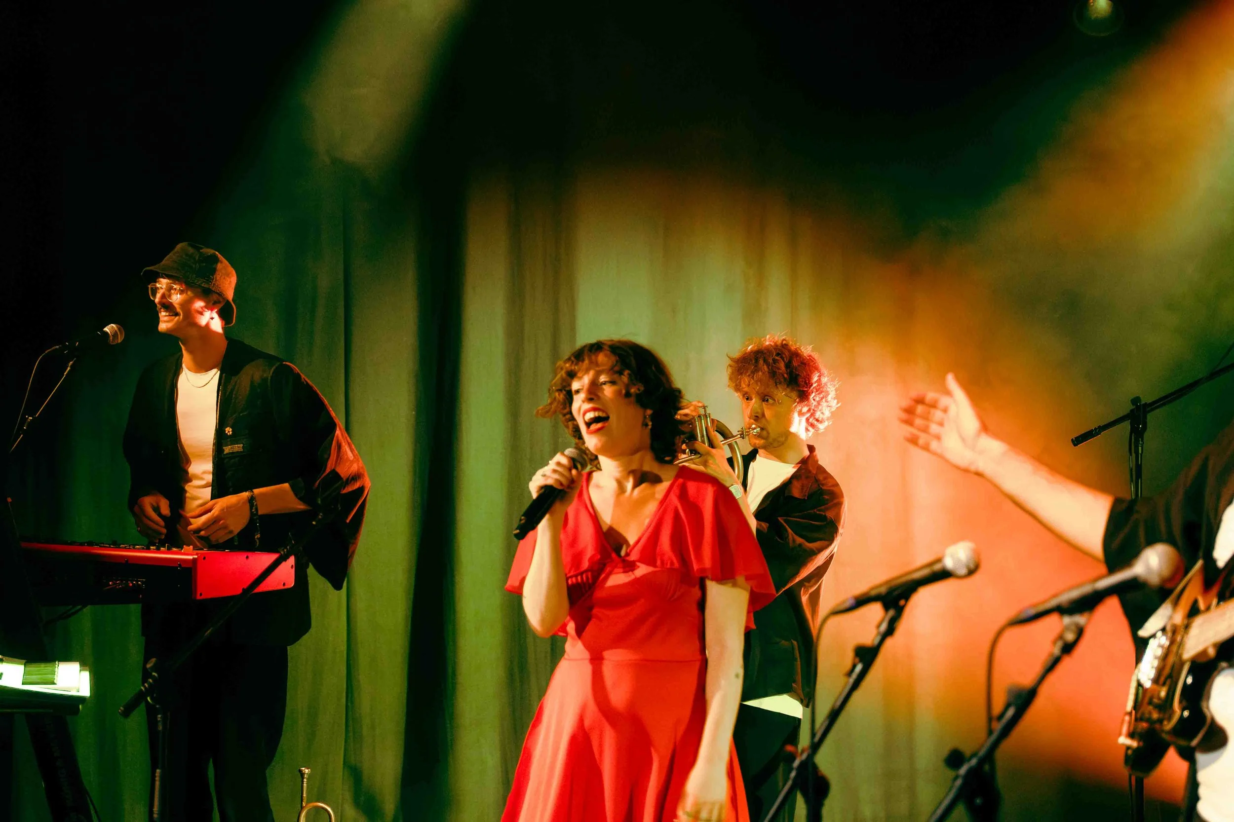 Een muzikale performance op een podium met drie muzikanten, waaronder een vrouw in een rode jurk die zingt en anderen die instrumenten bespelen, met gekleurde podiumverlichting.