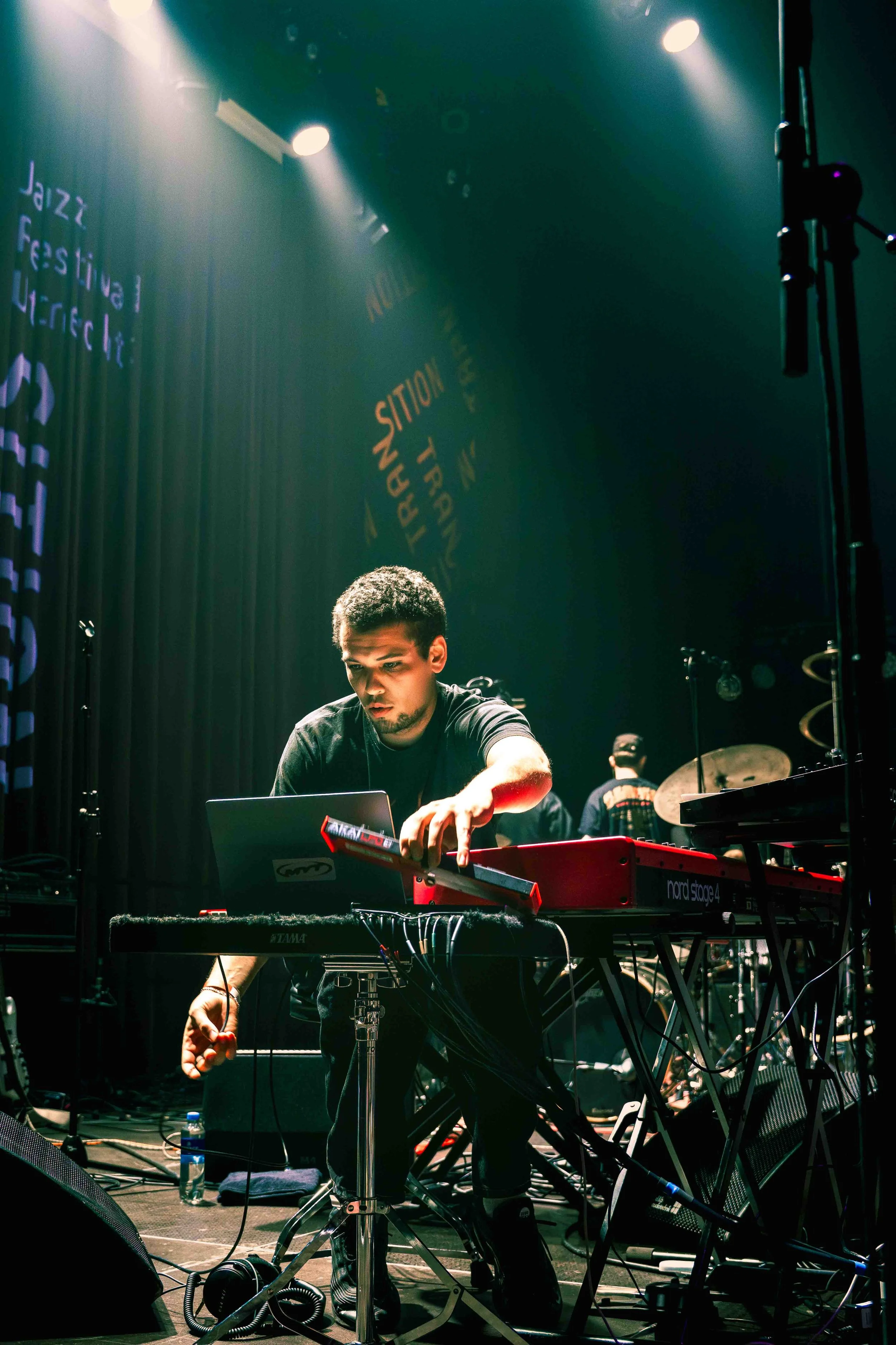 Een muzikant die live optreedt op een podium met synthesizers en andere elektronische apparatuur, met een achtergrond met woorden zoals 'Jazz Festival' en 'Transition'.