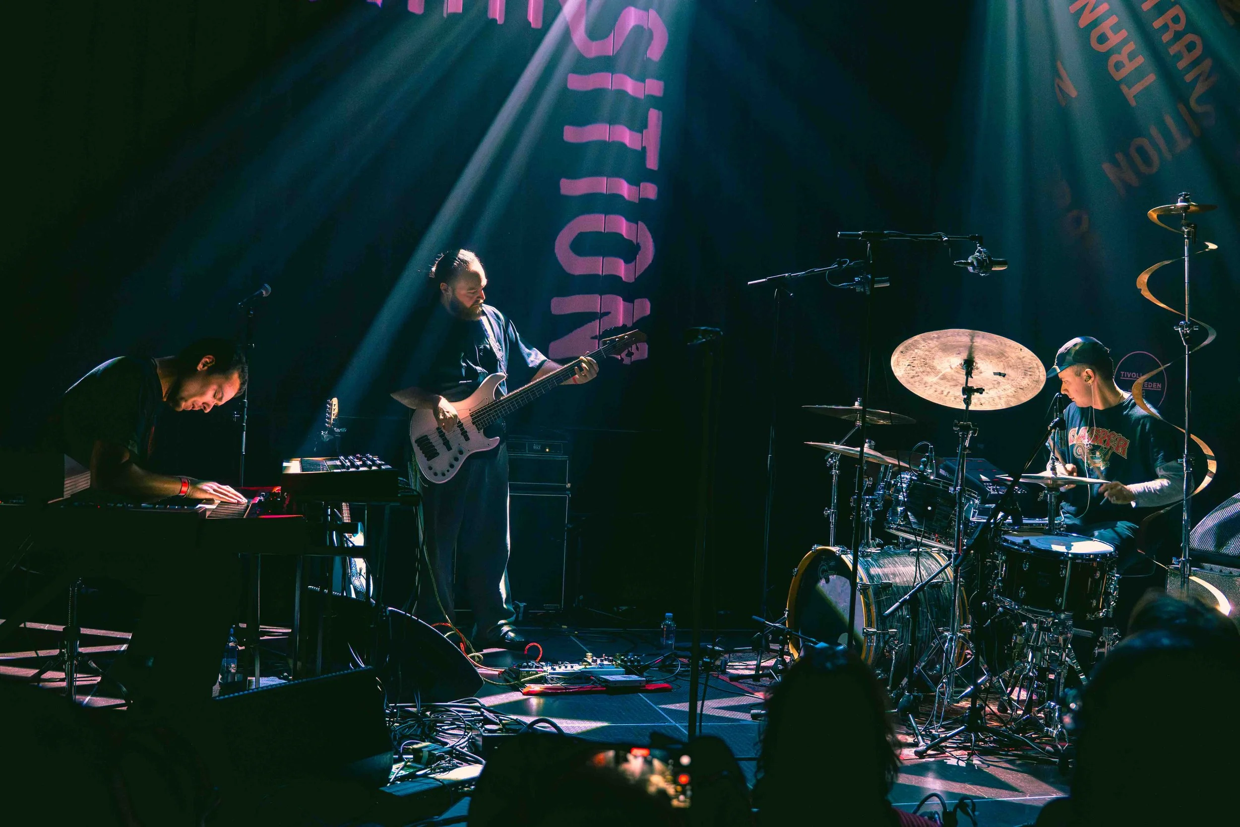 Een band met drie muzikanten op een podium, speel ze hun instrumenten. Eén persoon speelt keyboard, een ander speelt basgitaar, en de derde speelt drums. Er zijn verschillende microfoons en versterkers zichtbaar, met een achtergrondbanner en verlicht