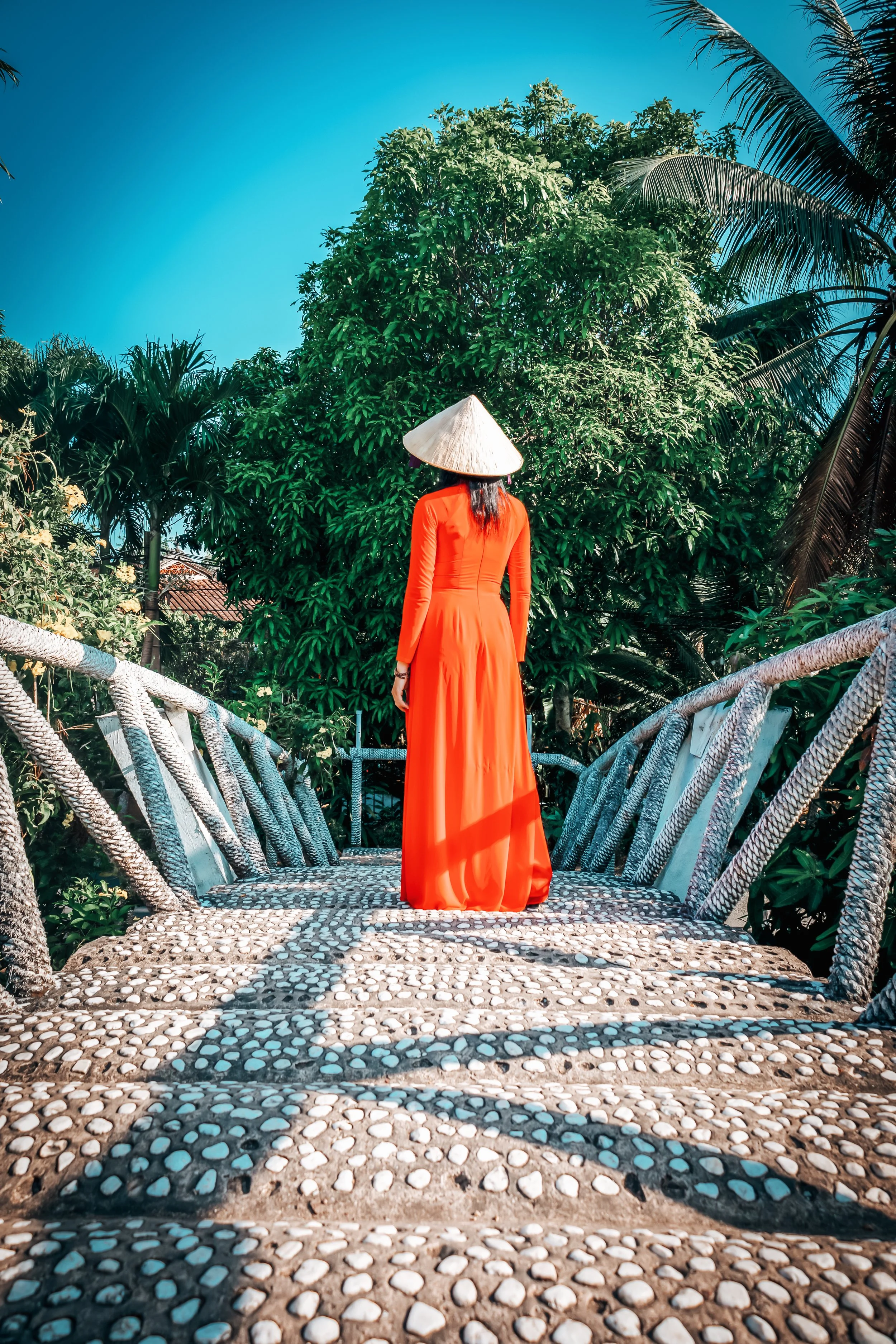 Femme en robe traditionnelle ao dai rouge et chapeau conique sur un pont de galets à Hội An, Vietnam