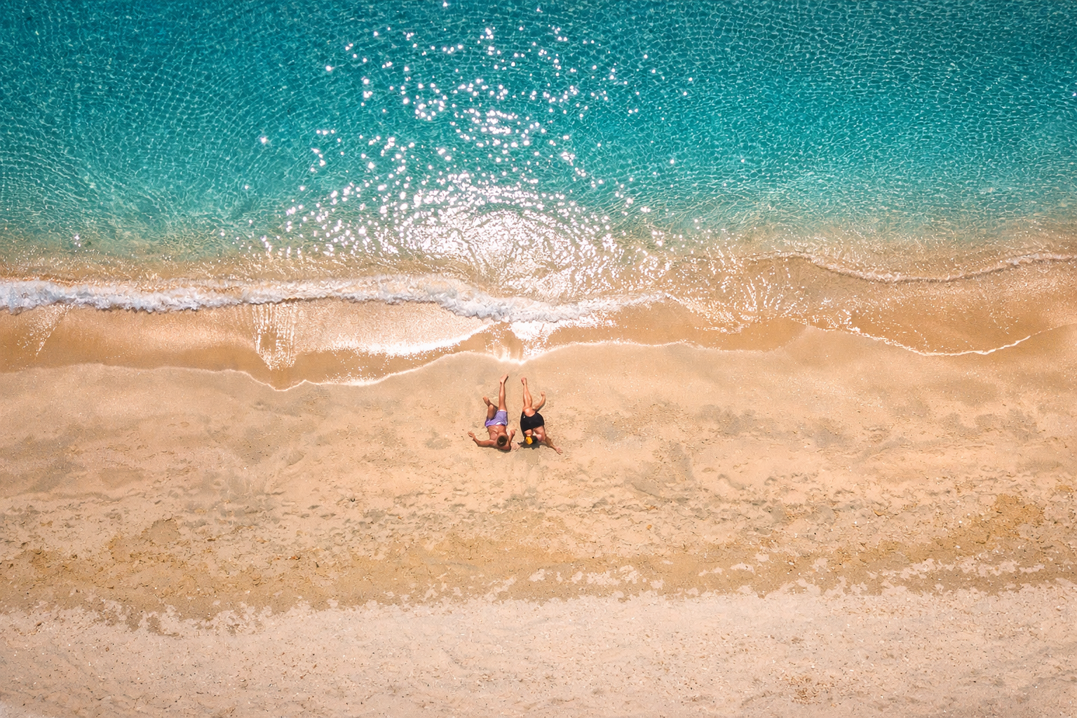 Vue aérienne de deux personnes allongées sur une plage de sable doré bordée d'eau turquoise scintillante, détente vacances tropicales