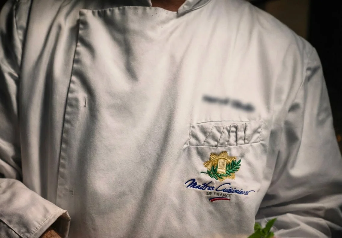  Gros plan sur veste blanche de chef brodée du logo Maître Cuisinier de France, excellence et savoir-faire culinaire français
