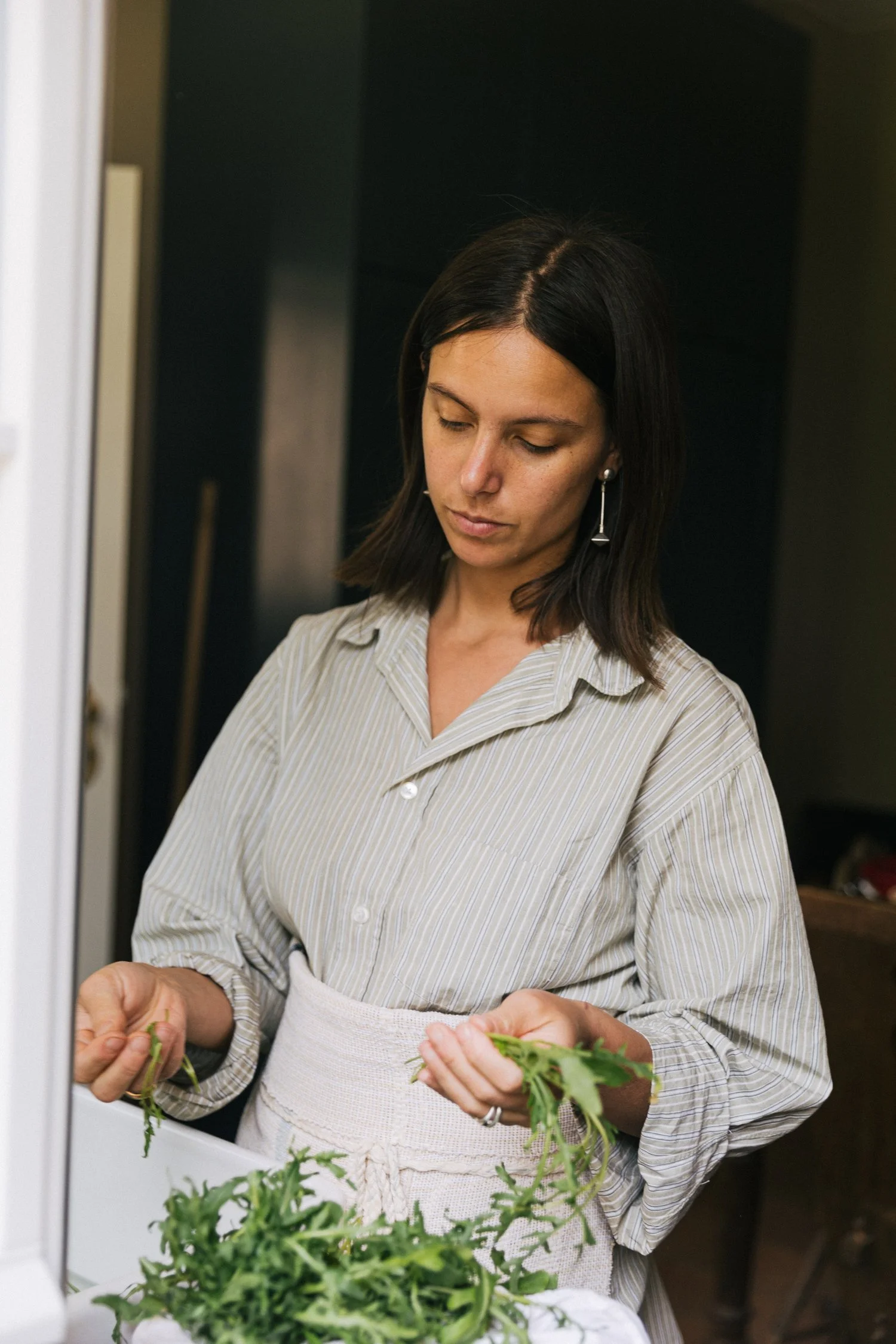 A woman with dark hair and a beige striped shirt is holding fresh green herbs in her hands while standing near a kitchen sink.