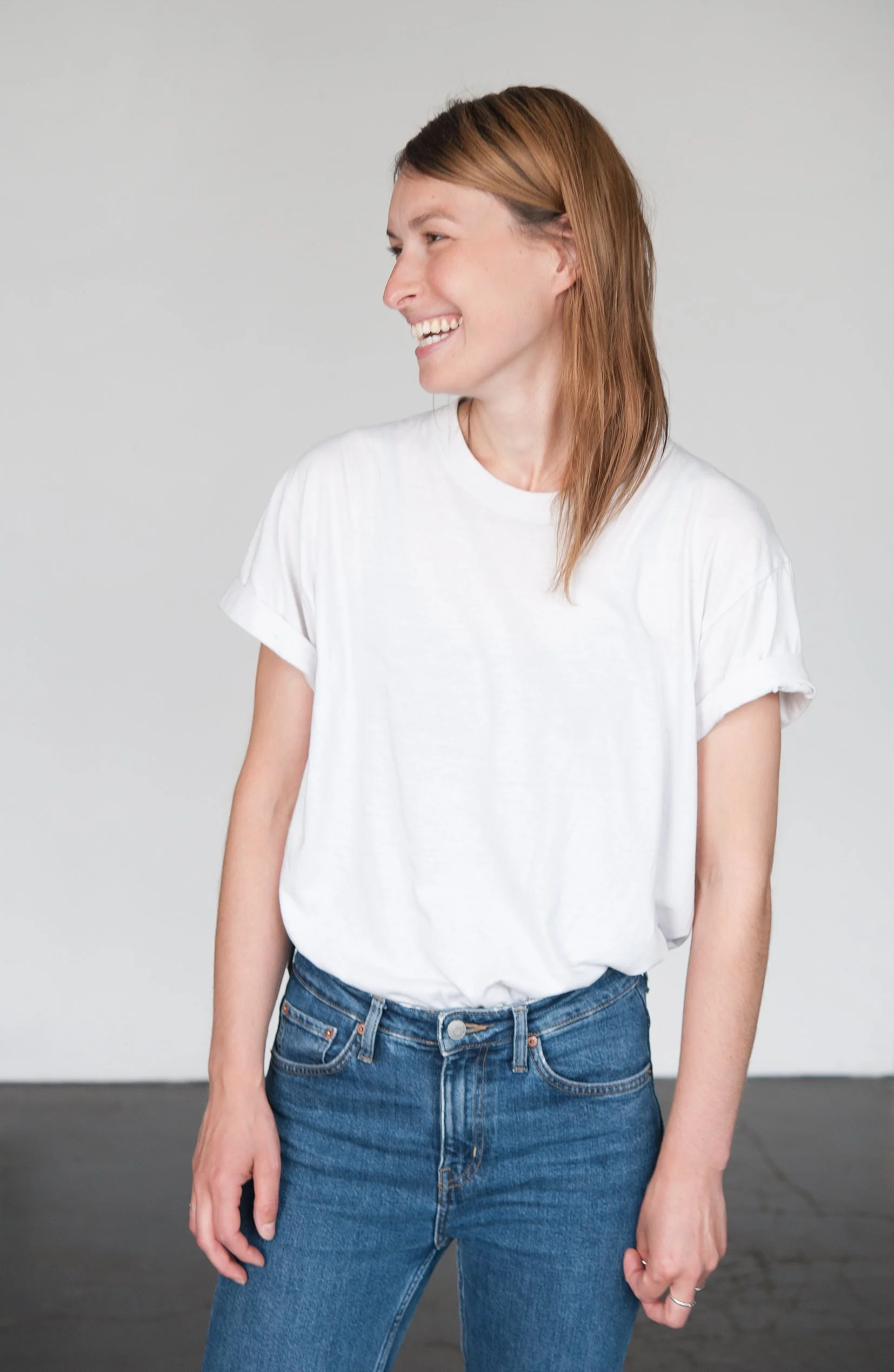 A woman with shoulder-length reddish-brown hair, smiling and looking to her left, standing against a plain light gray wall, wearing a loose white t-shirt and blue jeans.