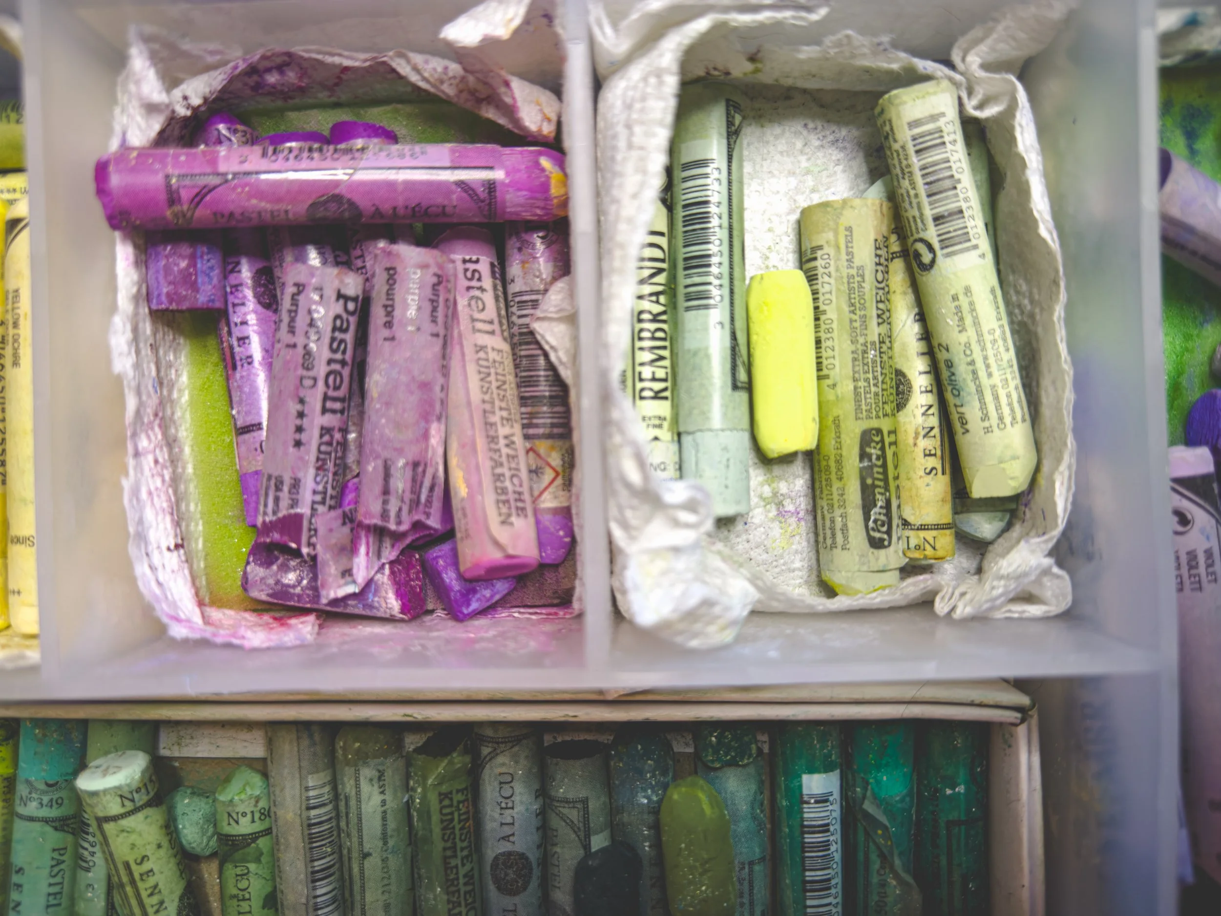 Dried pastel chalks arranged in separate compartments, with purple, yellow, and green colors, stored in a plastic container.