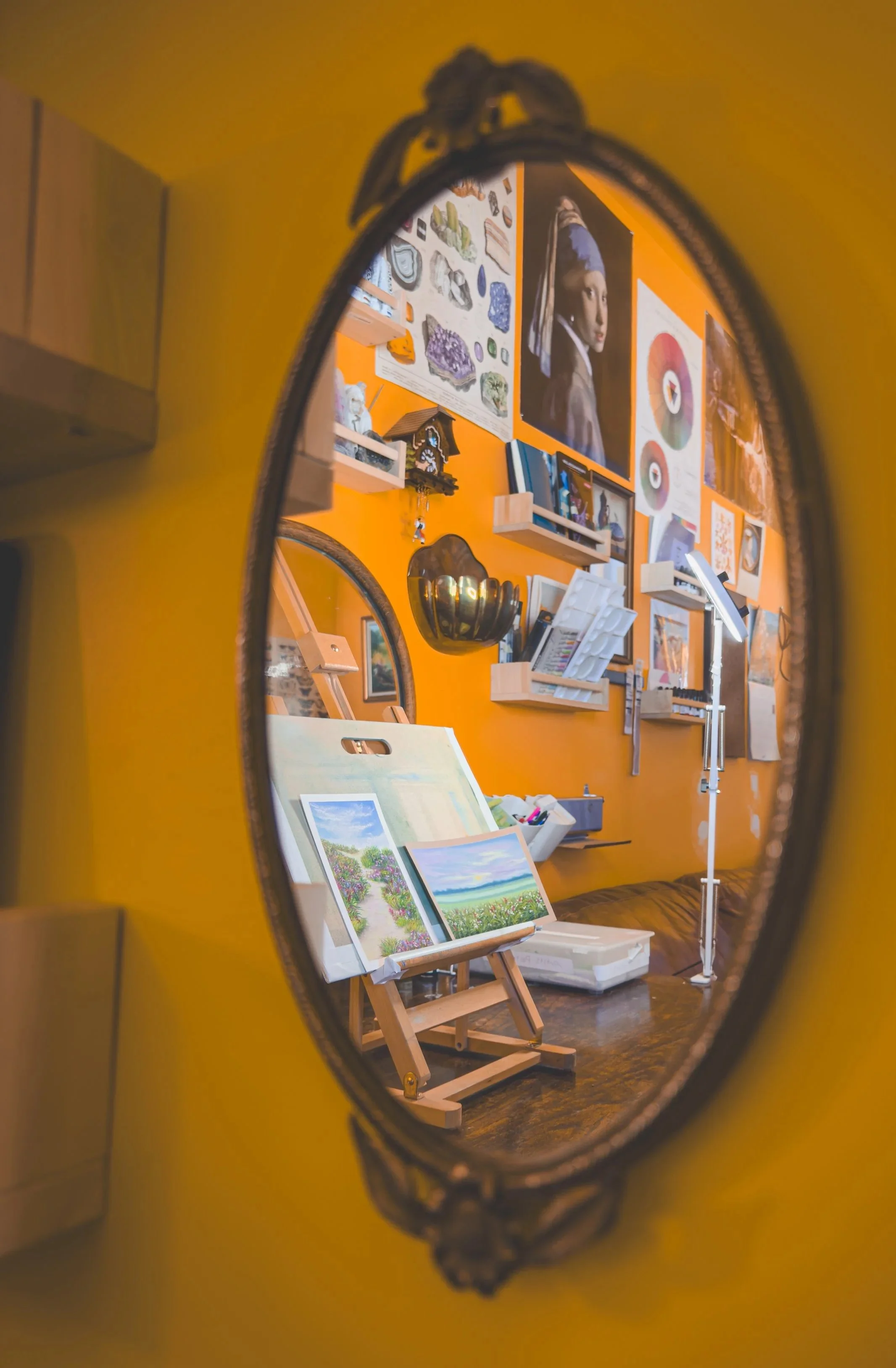 A mirror reflects an art studio with paintings, artwork, and art supplies on a vibrant orange wall.