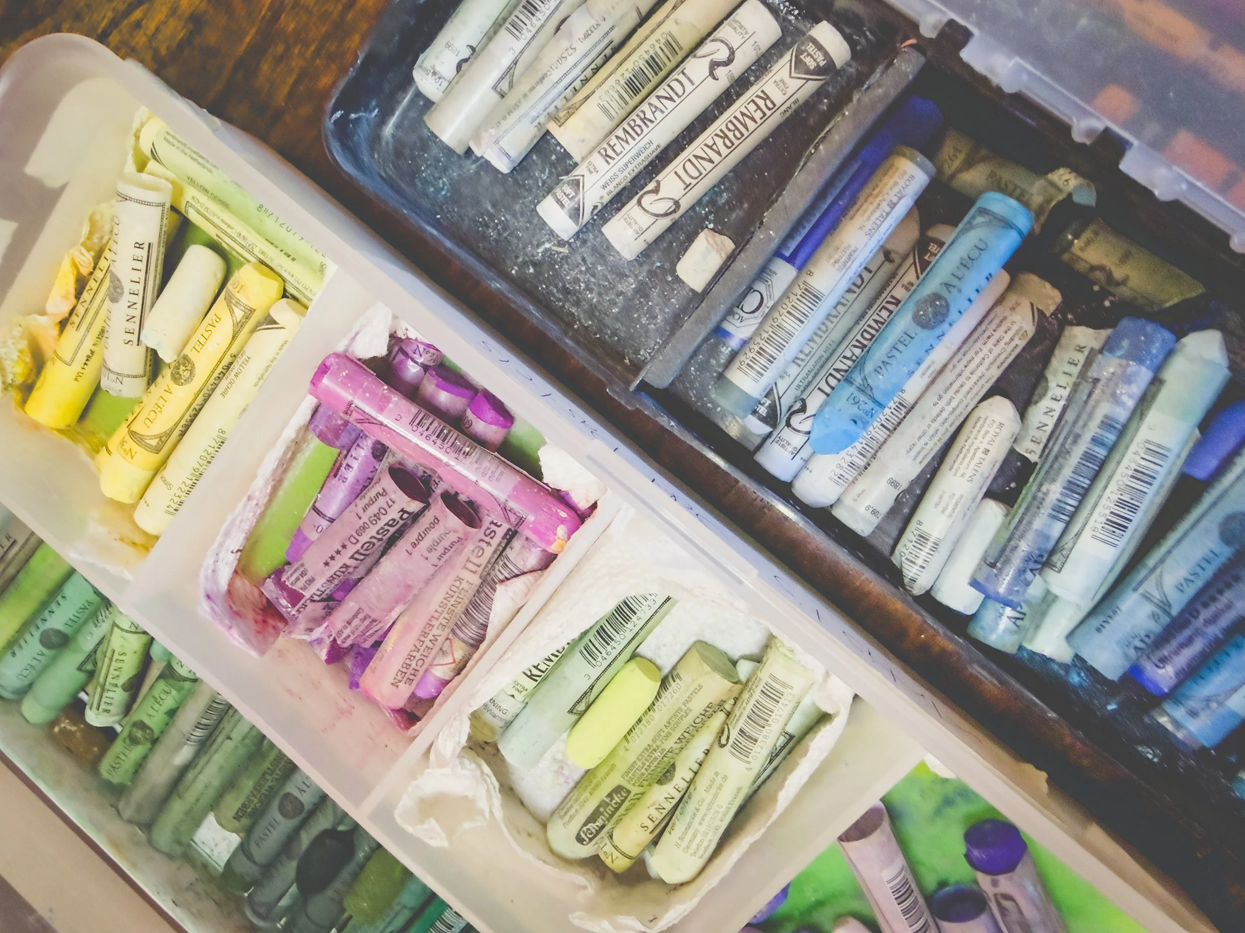 A collection of pastel chalks in various colors organized in separate containers, some with foil or paper wrappers, and a plastic lid with more chalks in the background.