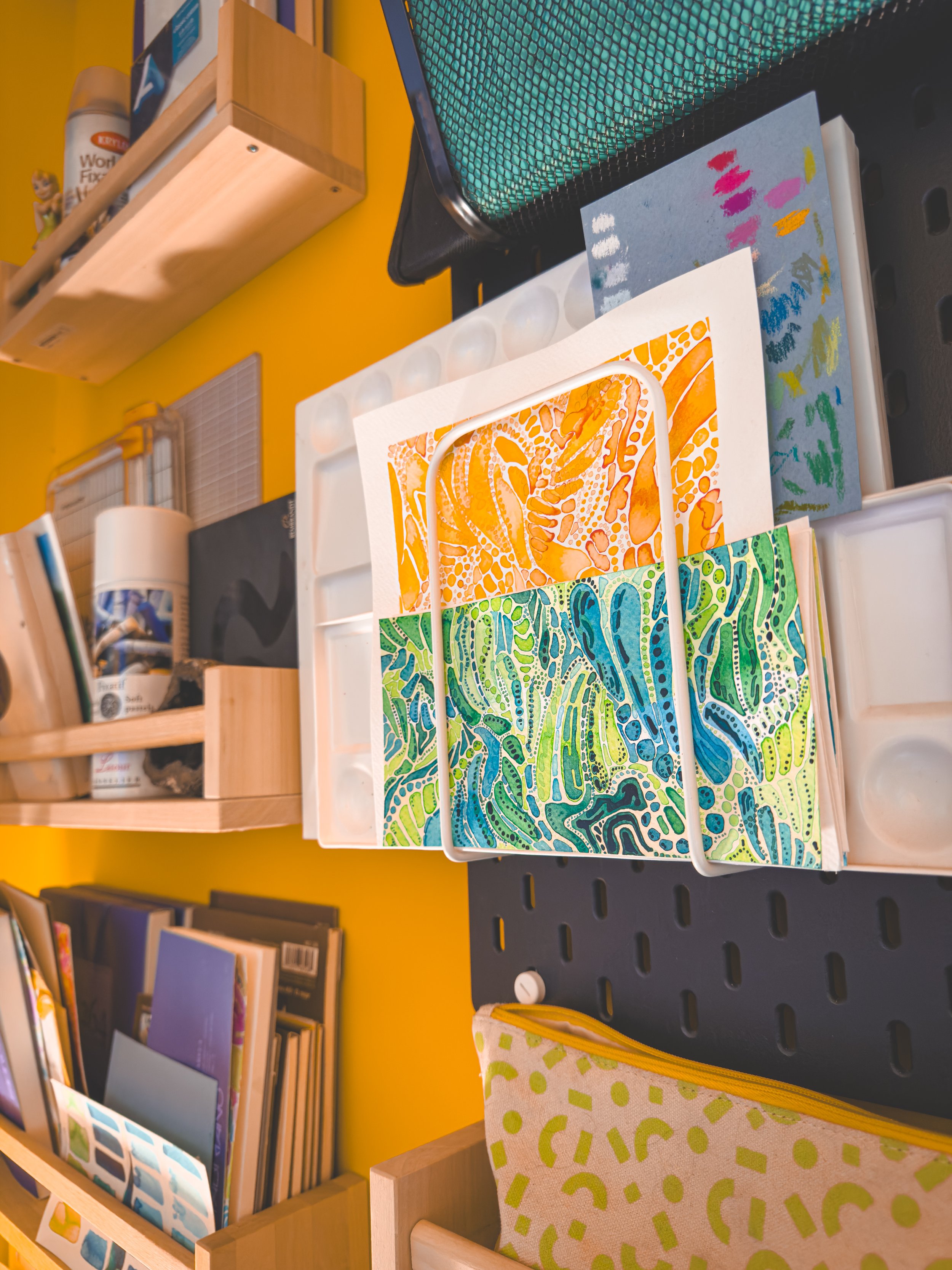 An art studio workspace with colorful abstract paintings, art supplies, books, and storage containers on shelves.
