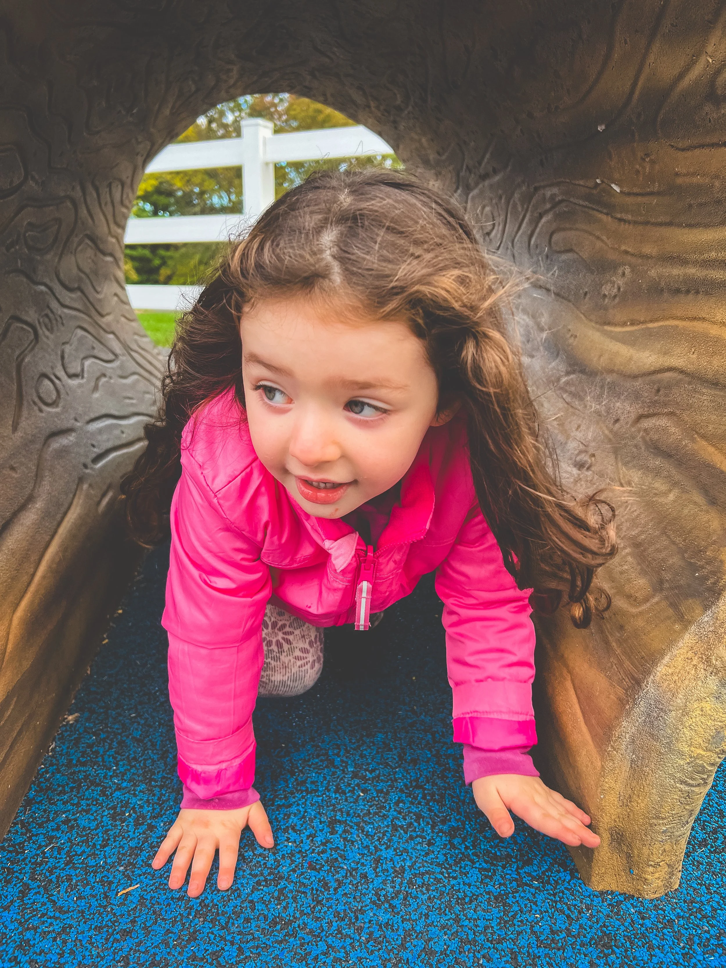 A young girl with brown hair wearing a pink jacket crawling through a playground tunnel.