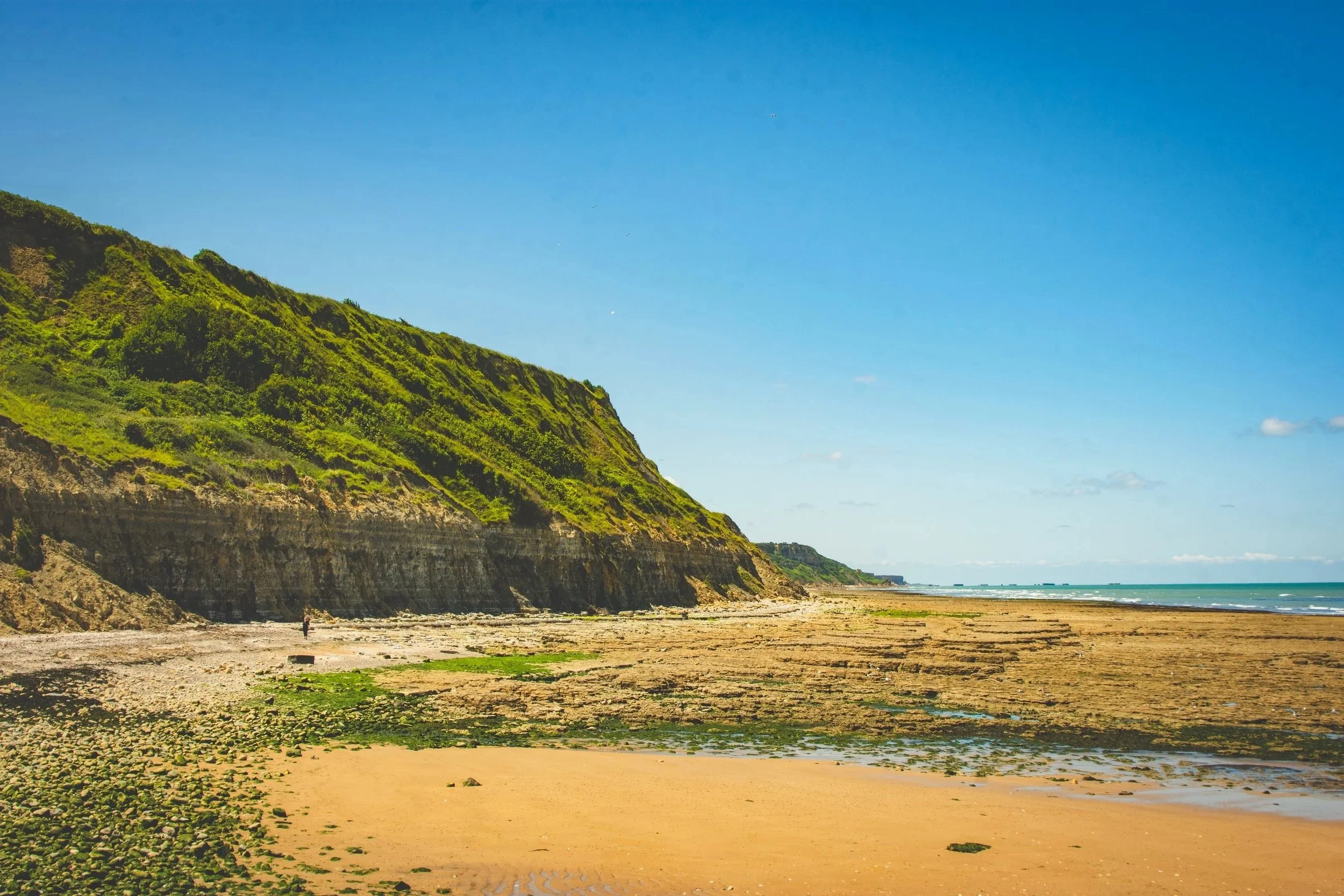 A sandy beach with green mossy rocks near cliffs on the French coast with lush green vegetation under a clear blue sky.