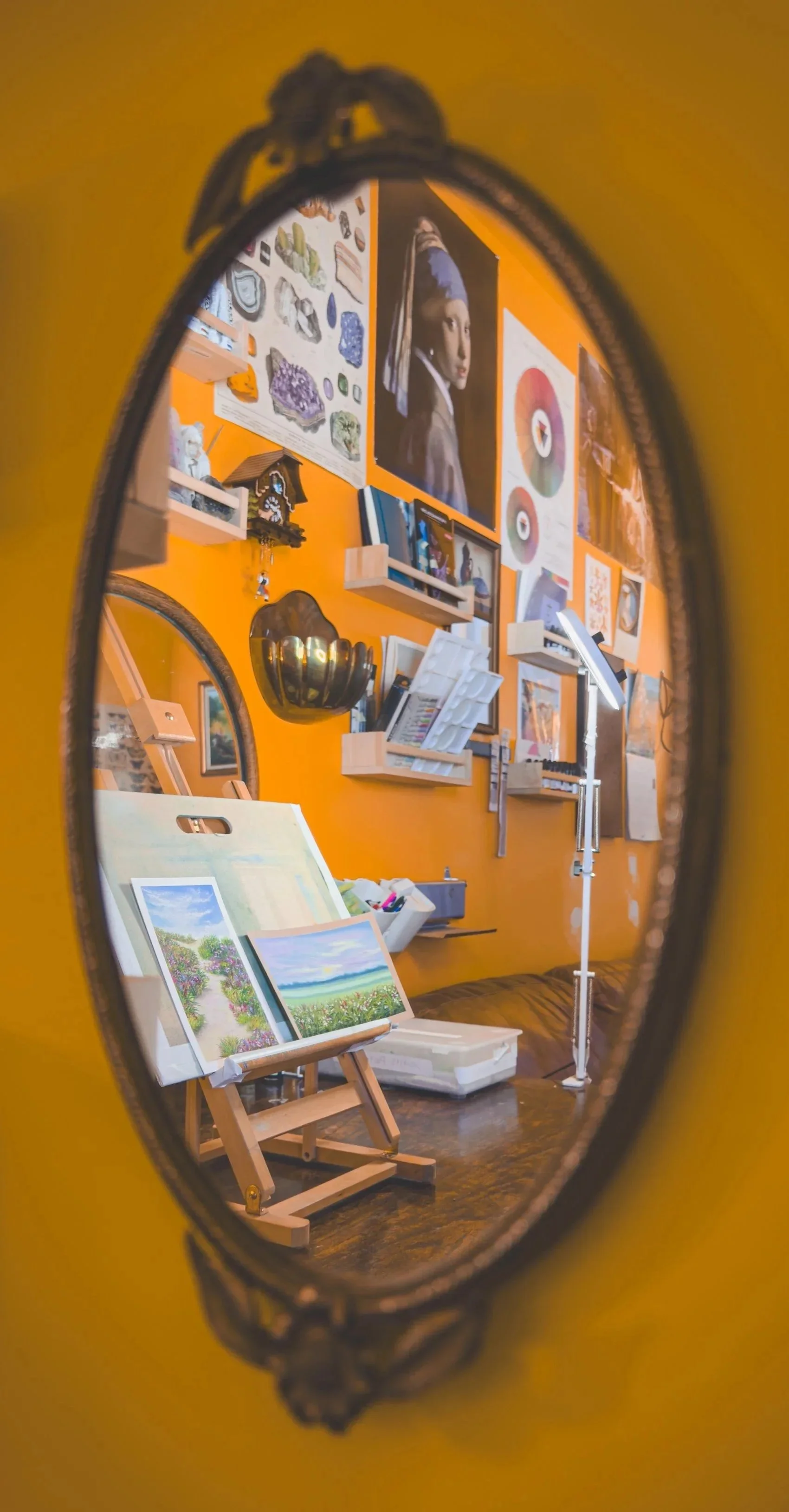 A reflection in an oval mirror shows an art studio with paintings, photographs, and art supplies on a bright orange wall.
