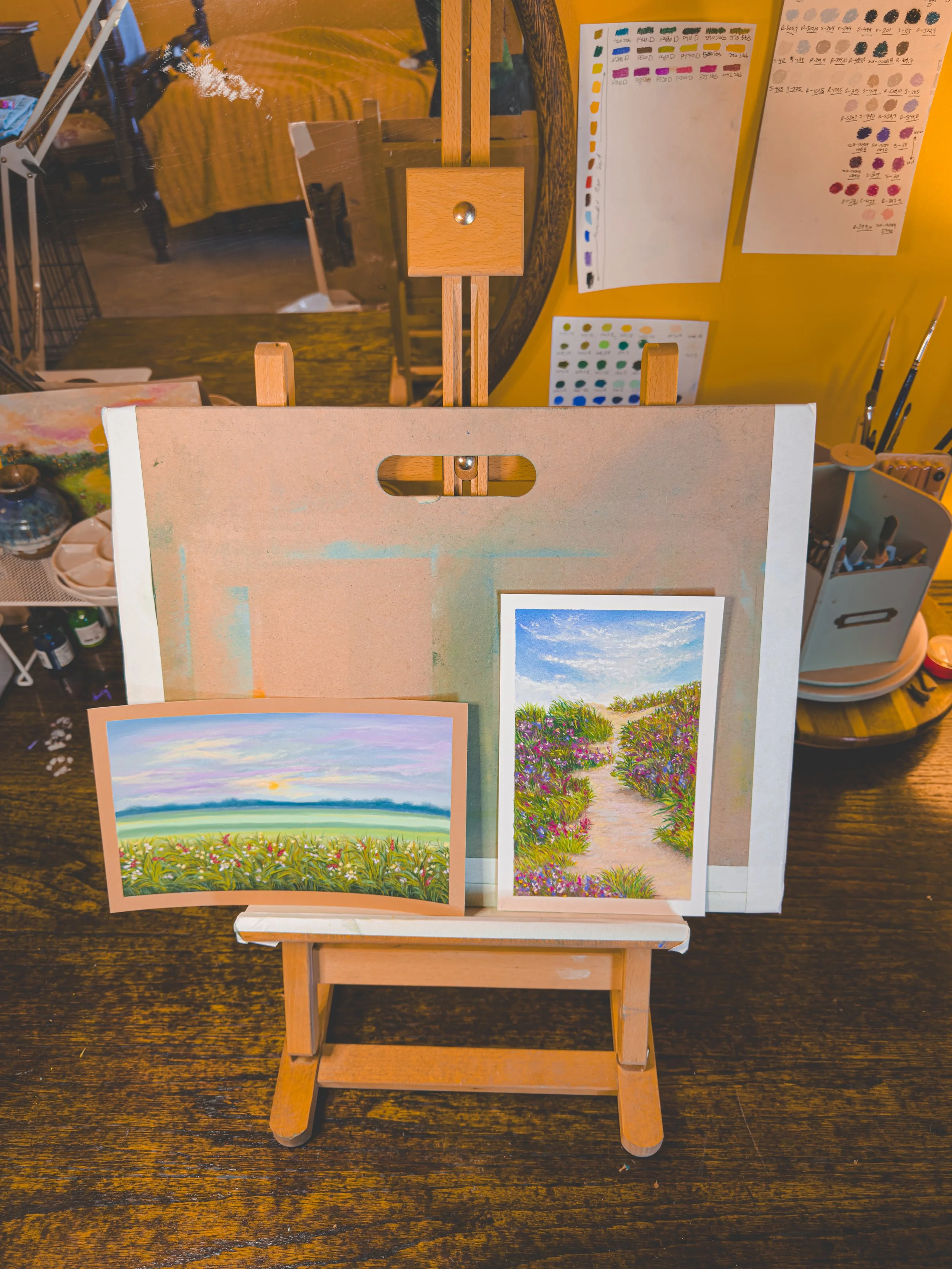 Two small landscape paintings on a miniature wooden easel, one depicting a sunset over a field, and the other showing a flower garden path, with a larger, blank canvas and art supplies in the background.