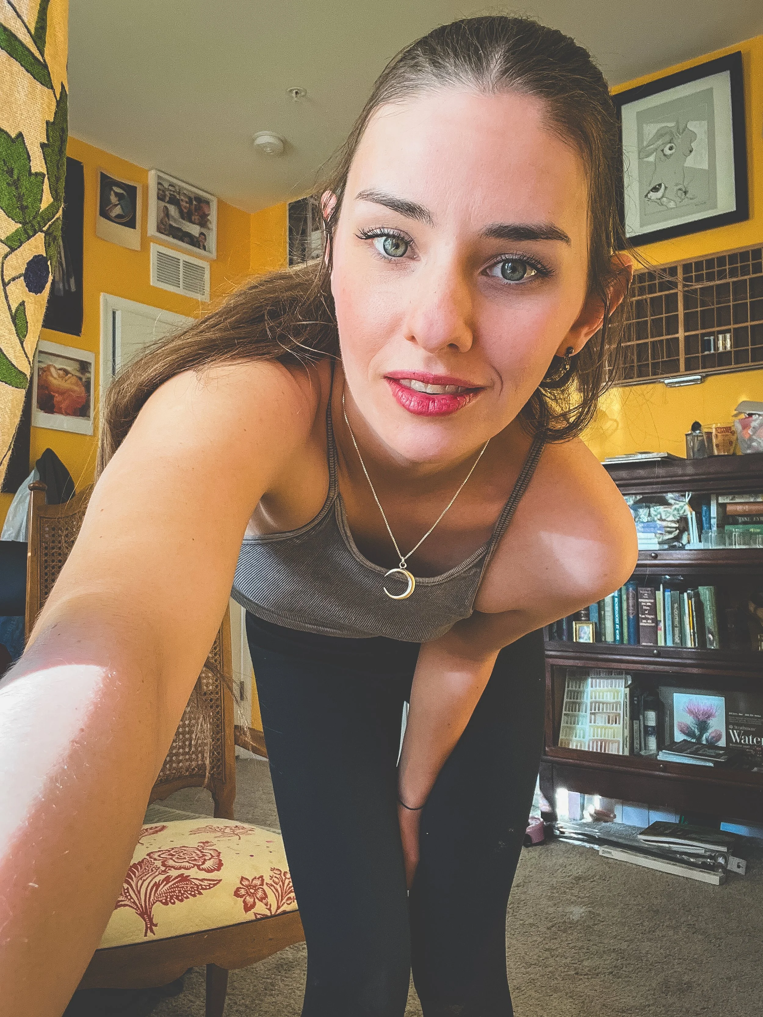 A woman with long brown hair, wearing a gray tank top and black pants, is leaning forward and smiling at the camera inside a colorful room with yellow walls, framed artwork, and a bookcase.