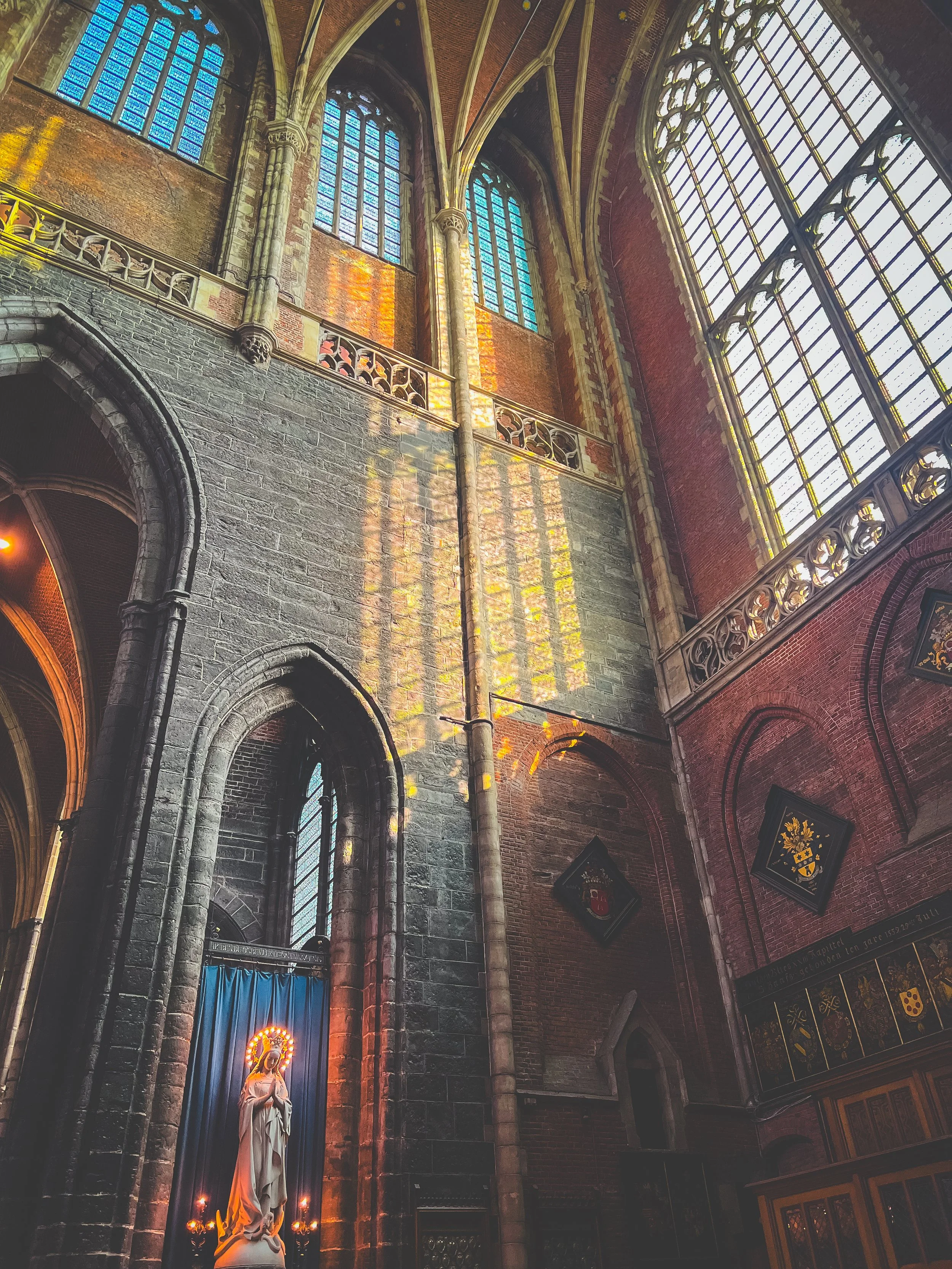 Sunlight streams through tall stained glass windows illuminating the interior of a Gothic-style church with stone arches and walls, a statue of the Virgin Mary with candles, and decorative shields and plaques on the walls. Ghent cathedral.