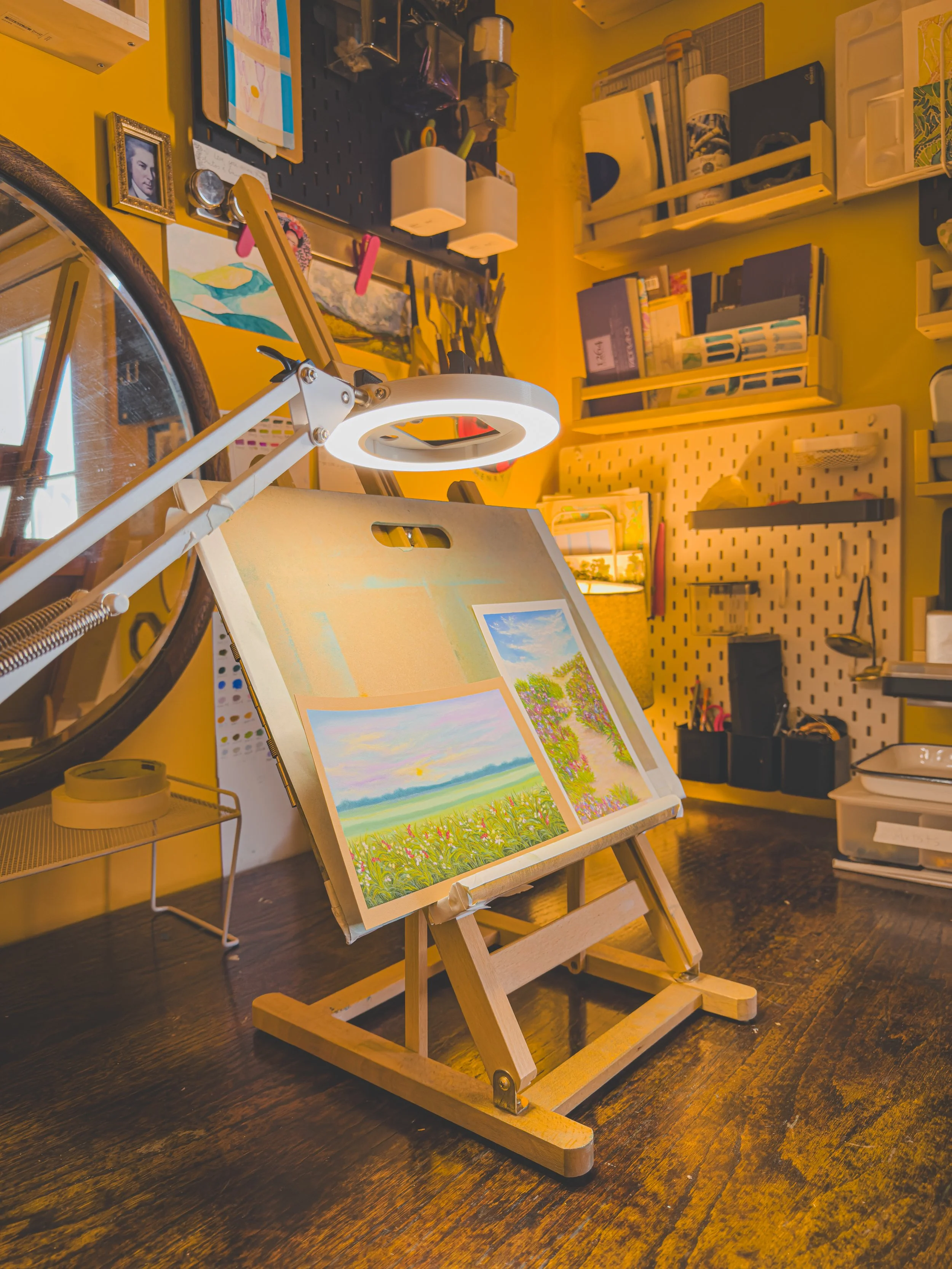 An art workspace with paintings on a small wooden easel, bright yellow walls, and various art supplies and storage shelves in the background.