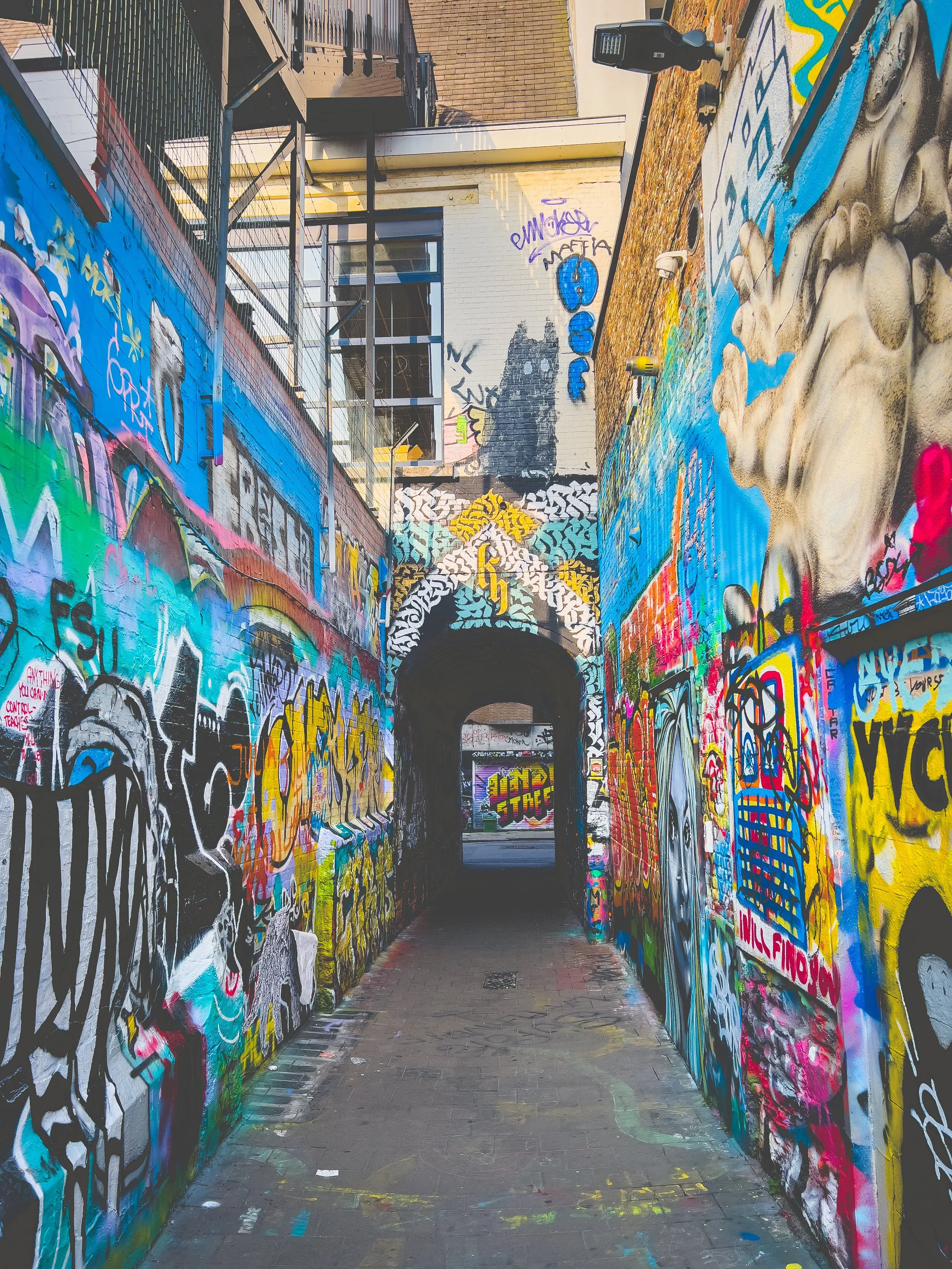An alleyway in Ghent covered in colorful graffiti art, leading to a dark passageway with an arch, surrounded by buildings with large windows and a fire escape.