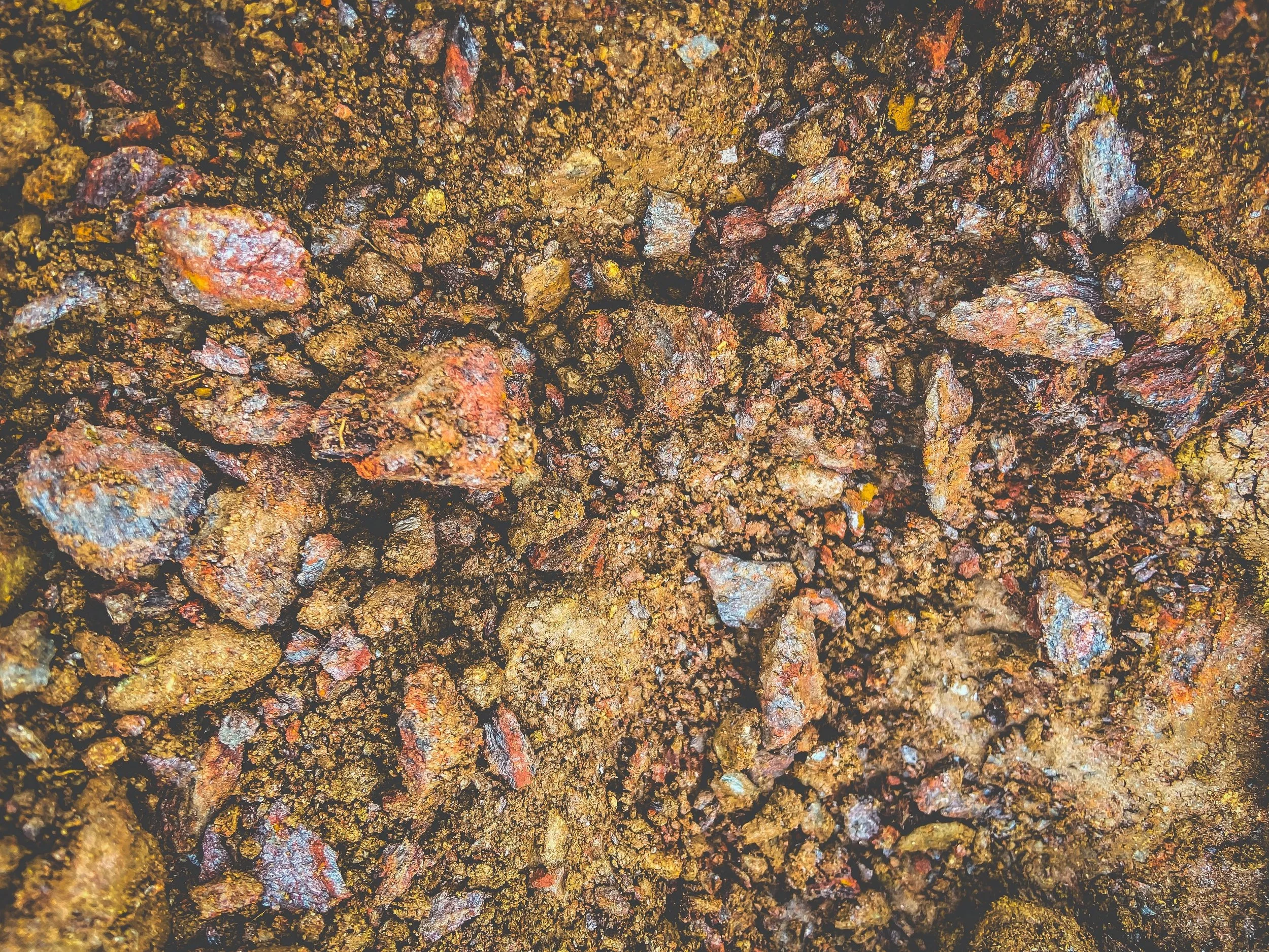Close-up of a textured soil surface with small rocks and dirt, showing various shades of brown, red, and yellow.