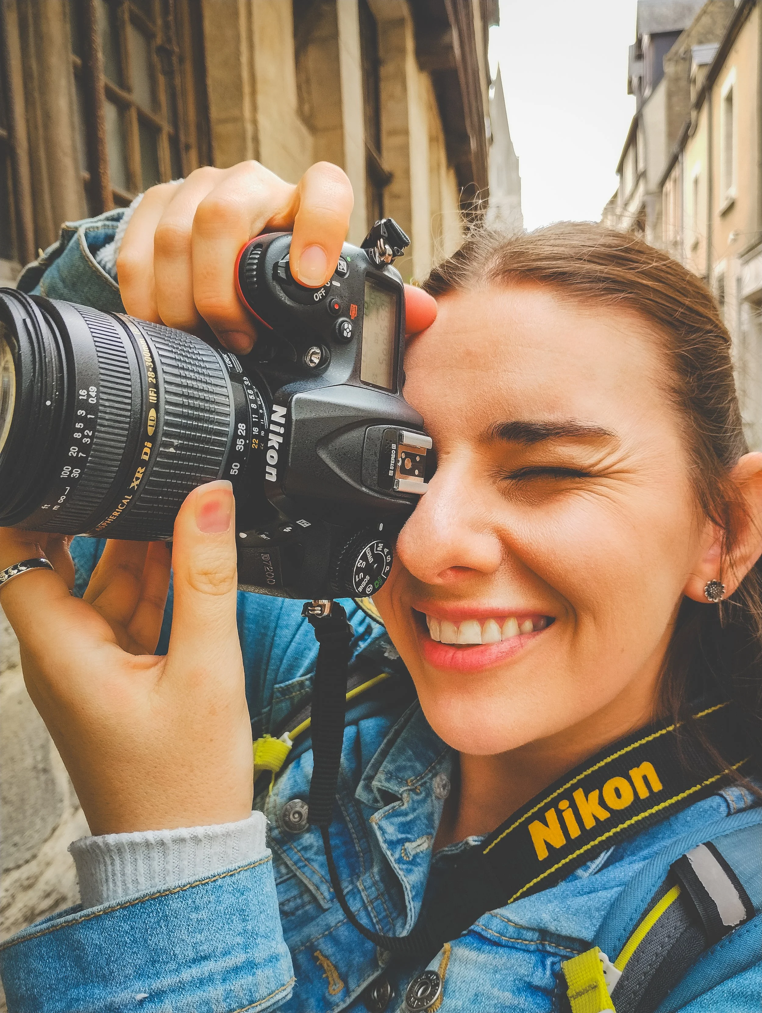 A woman smiling while looking through a Nikon d7200 camera's viewfinder on a street in France with old stone buildings.