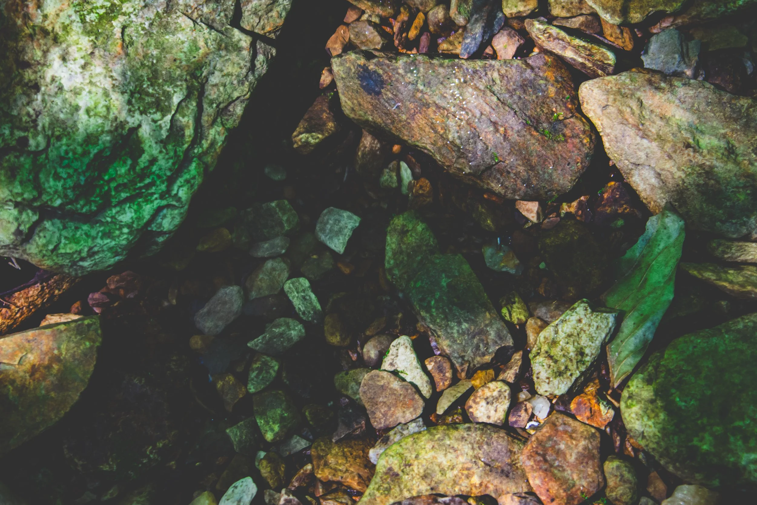 Close-up of rocks and pebbles in a natural stream or riverbed, with some covered in green moss and with water visible in the gaps.