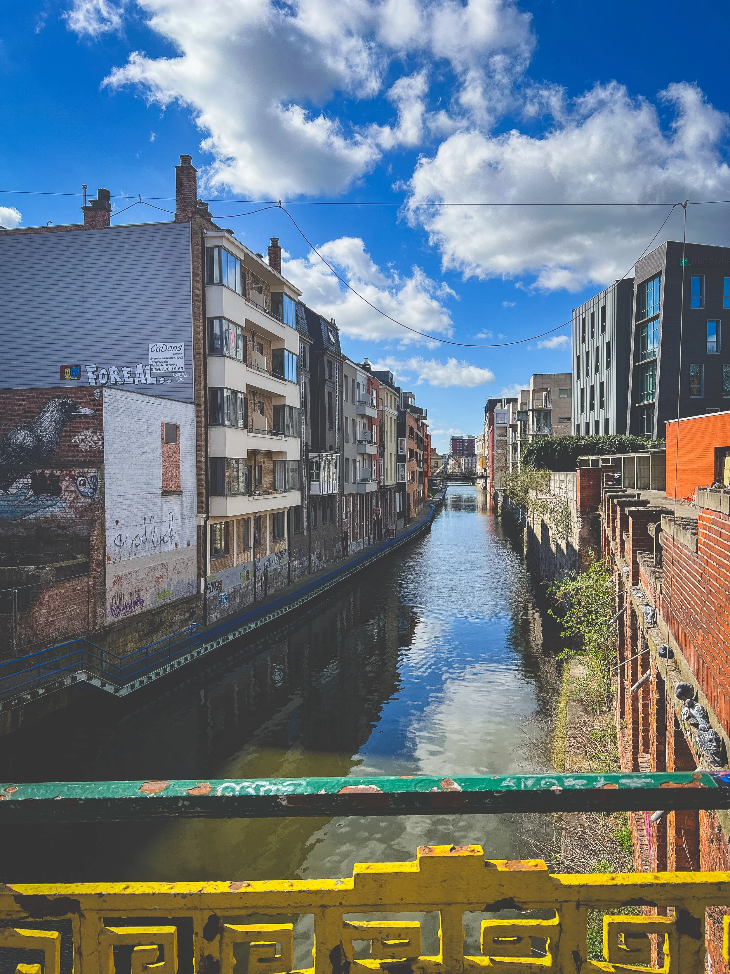 A city canal lined with modern apartment buildings under a partly cloudy sky, with a yellow and green railing in the foreground. Gent, Belgium.