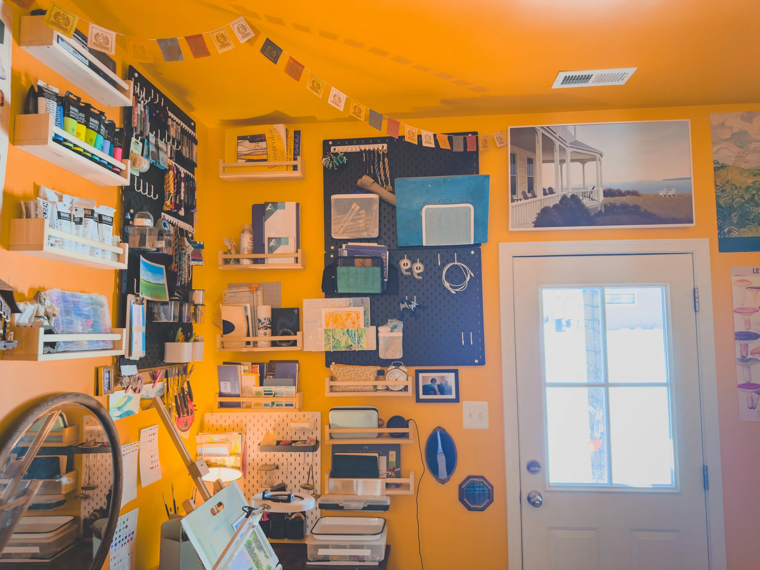 A brightly lit craft room with yellow walls decorated with artwork and decorations, featuring organized shelves with art supplies and tools, a large window and door providing natural light.