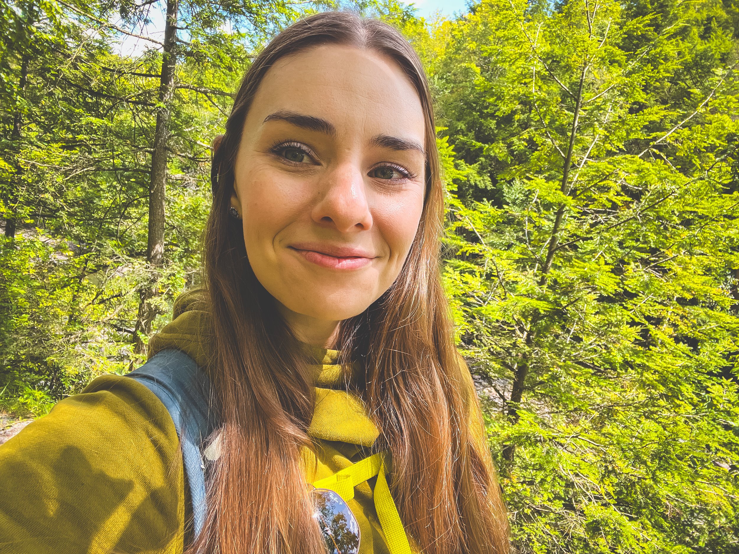 A young woman with long brown hair and green eyes taking a selfie outdoors in a wooded area, wearing an olive jacket and a blue backpack.