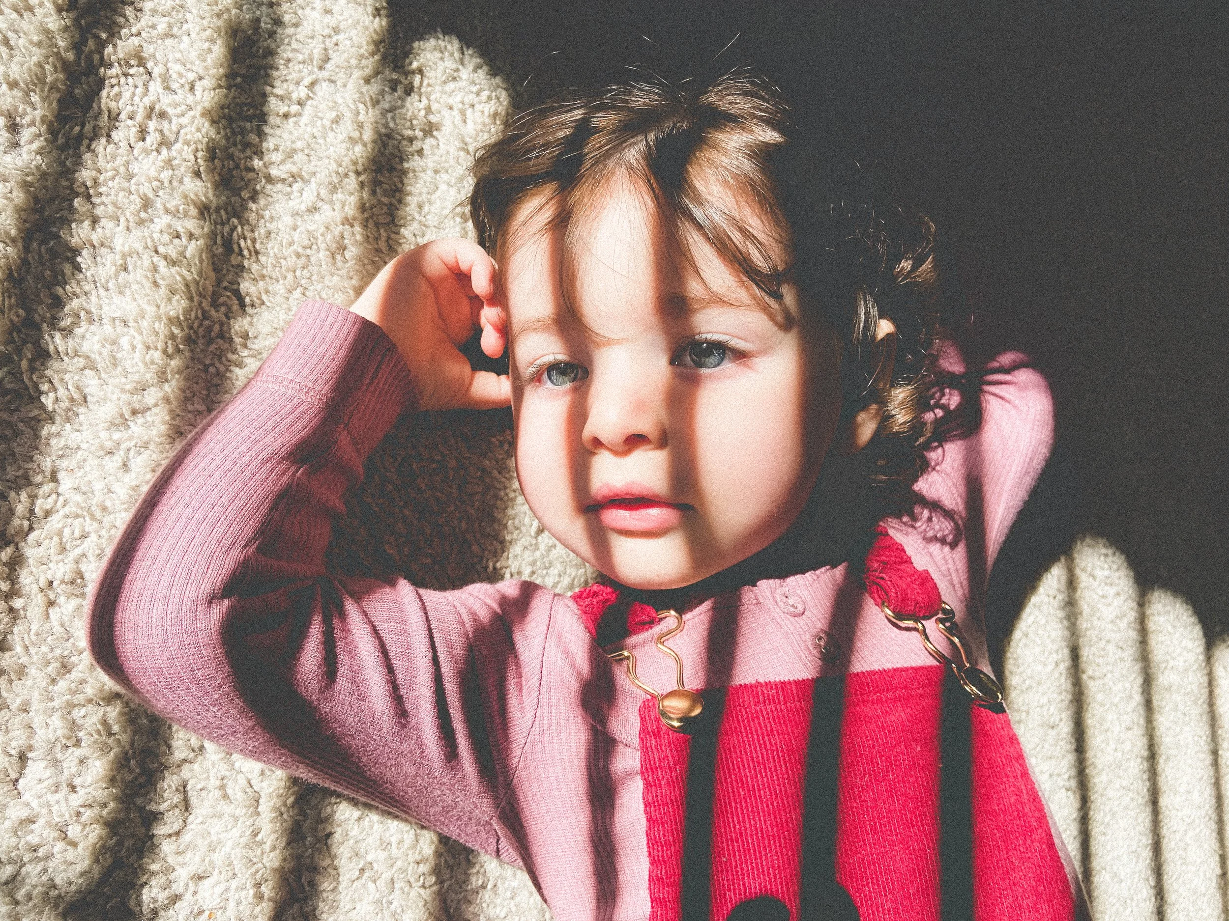 A young girl with curly brown hair and blue eyes lying on her back on a soft, beige surface, resting her head on her left arm, with sunlight casting shadows across her face.