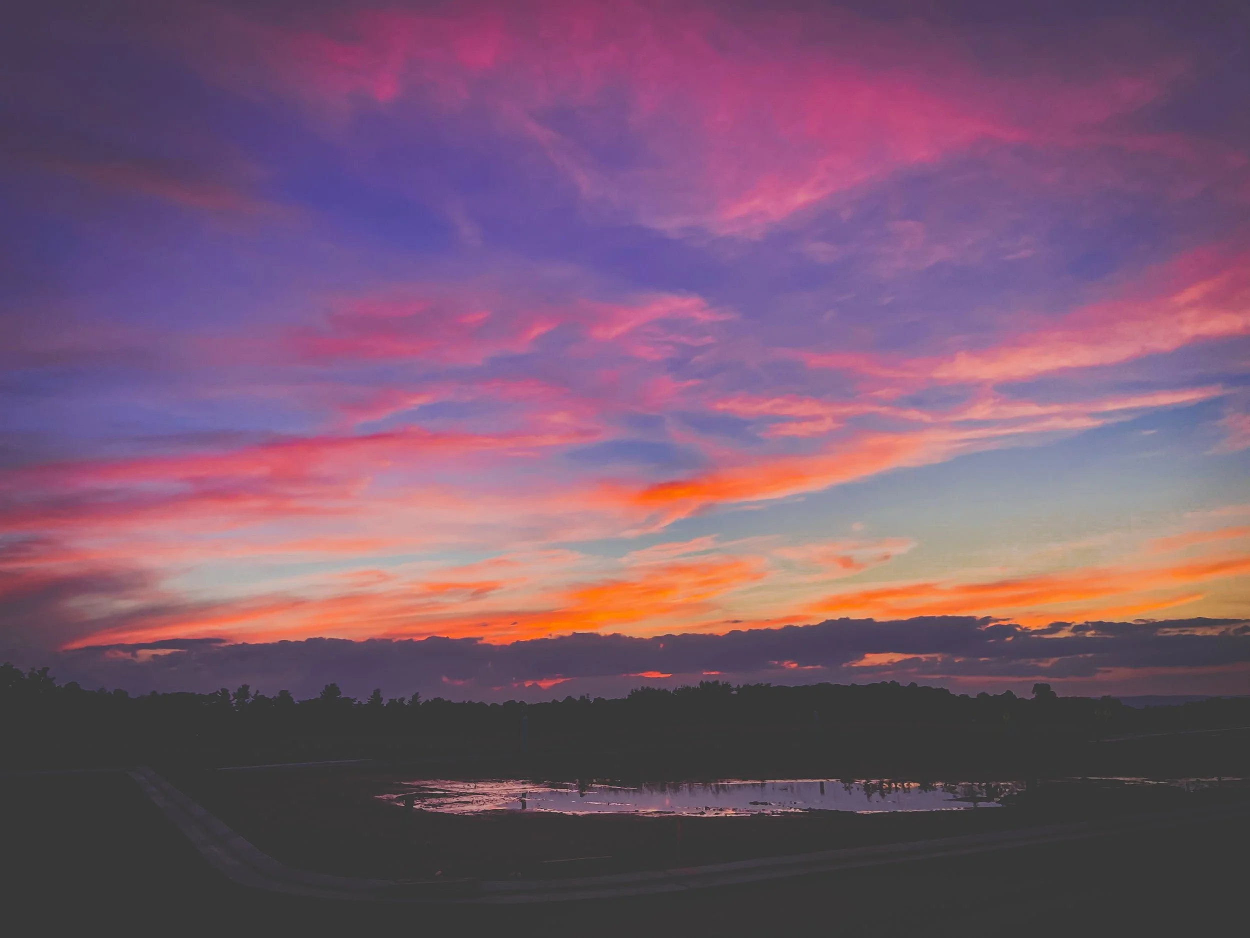 Colorful sunset sky with pink, purple, orange, and blue clouds over a silhouette of trees and a pond reflecting the sky.