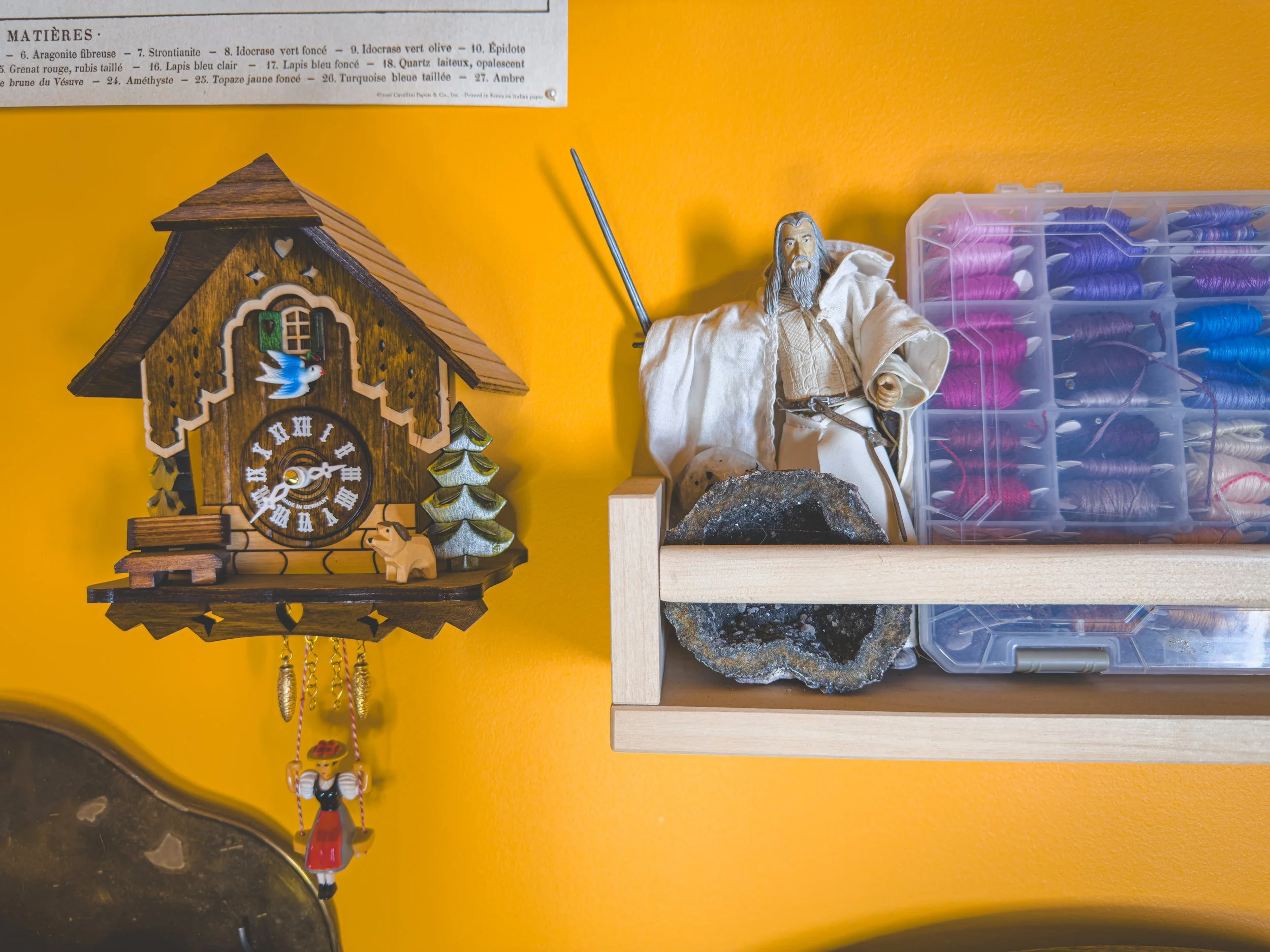 A wooden cuckoo clock with a blue bird, green window, and small figurines, hanging on a yellow wall. To the right, a shelf holds a detailed Gandalf figurine with a staff, a geode, and a storage container filled with various colored threads.
