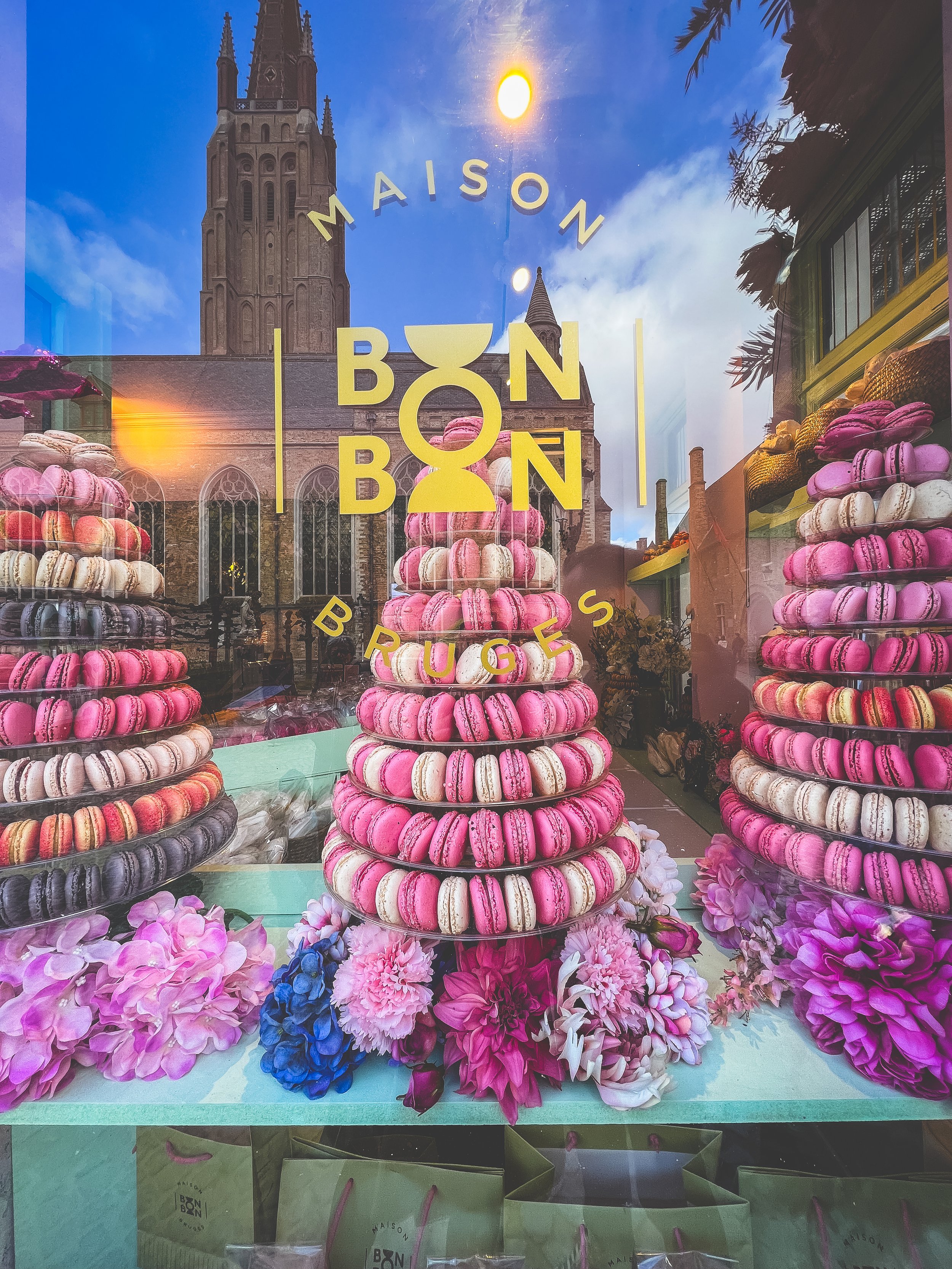 Display of pink, purple, beige, and black macarons arranged in tiered stacks on glass plates in a bakery shop window with the words 'MAISON BONBON BRUGES' on the glass, reflecting a church with tall spires and a blue sky.