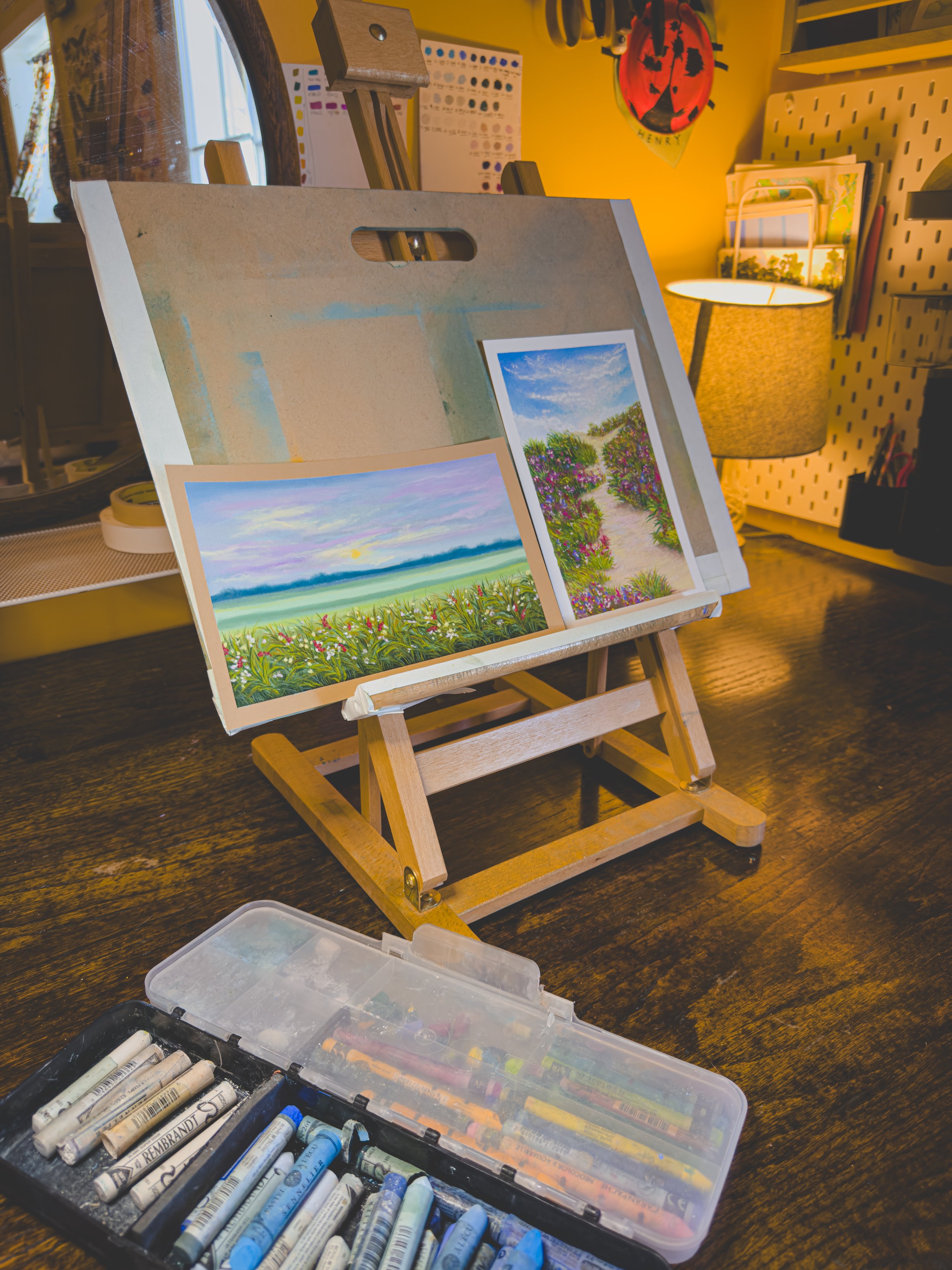 Two landscape paintings displayed on a small wooden easel, with a box of pastel sticks in the foreground, in a cozy room with warm lighting and decorative items on the wall.