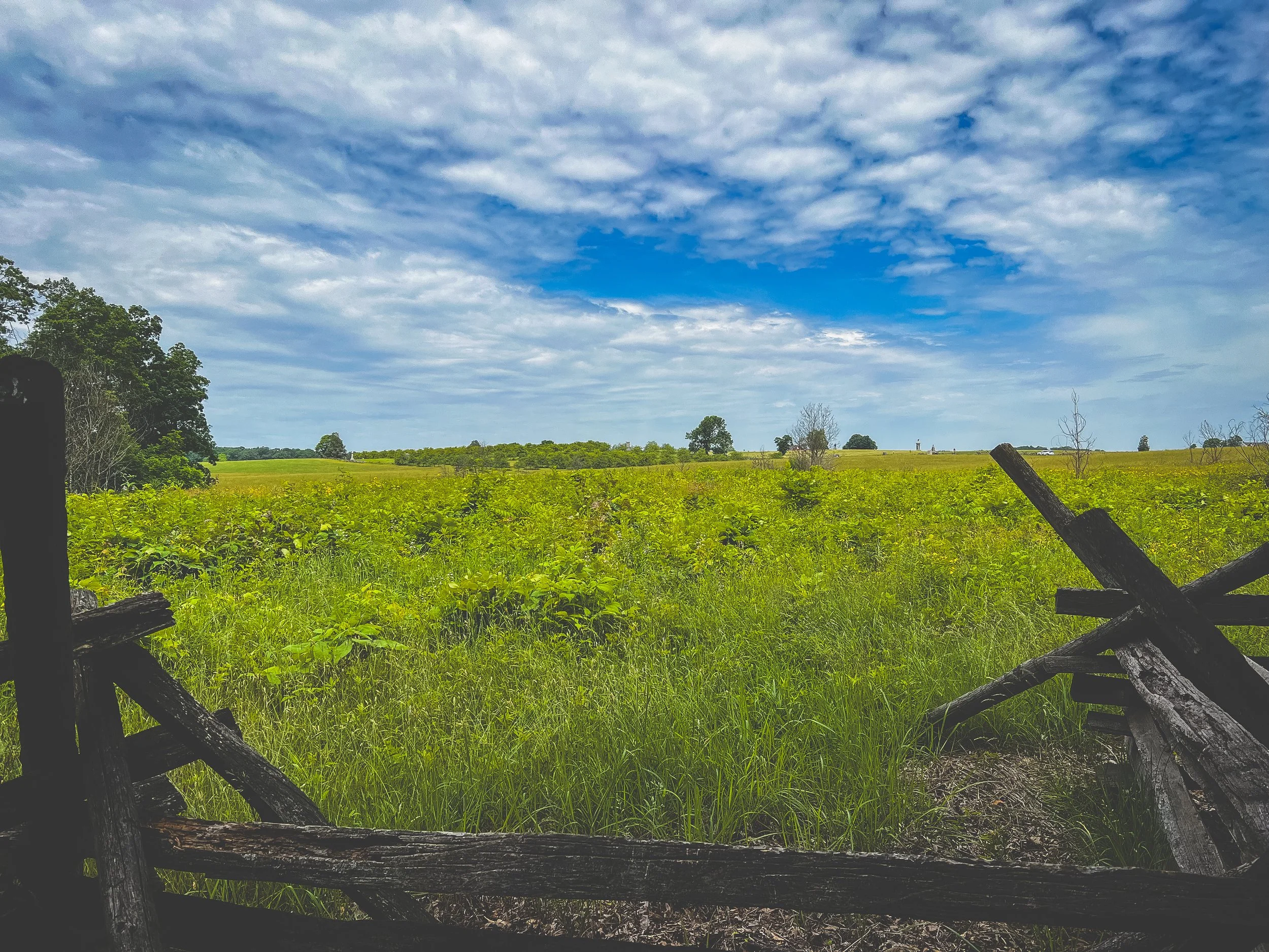 Open grassy field with scattered trees under a partly cloudy sky, viewed through a broken wooden fence, Gettysburg, Pennsylvania. 