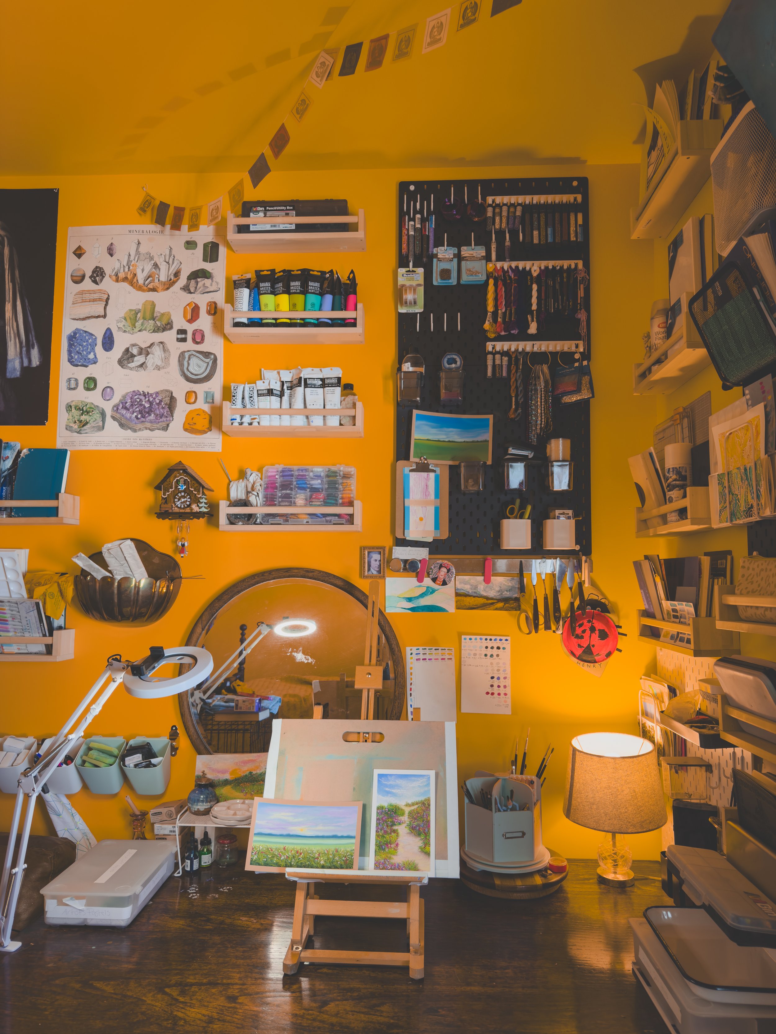 An art studio with a yellow wall, showing shelves with paint supplies, hanging hooks with tools, paintings, and a table with art supplies and paintings in progress, lit by a desk lamp and a table lamp.