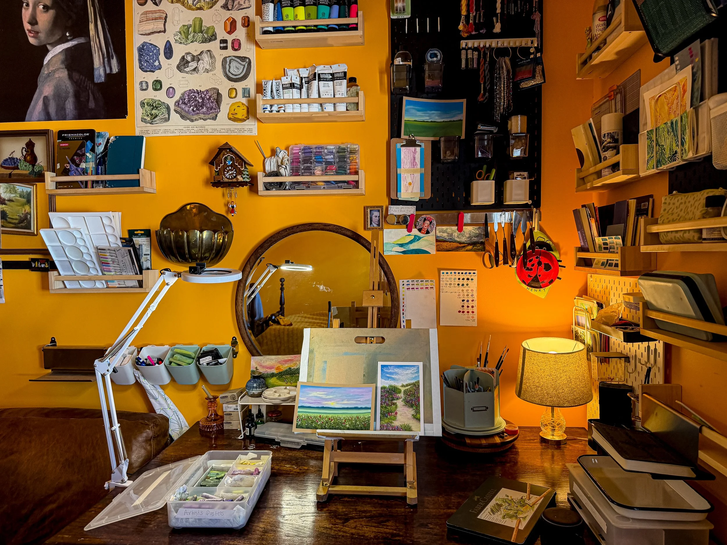 A cozy art and craft workspace with a yellow wall decorated with art supplies, framed artwork, and various tools. A wooden table holds paintings, a lamp, and art supplies. Shelves on the wall contain paint, brushes, books, and decorative items. A round mirror hangs on the wall, reflecting part of the room.