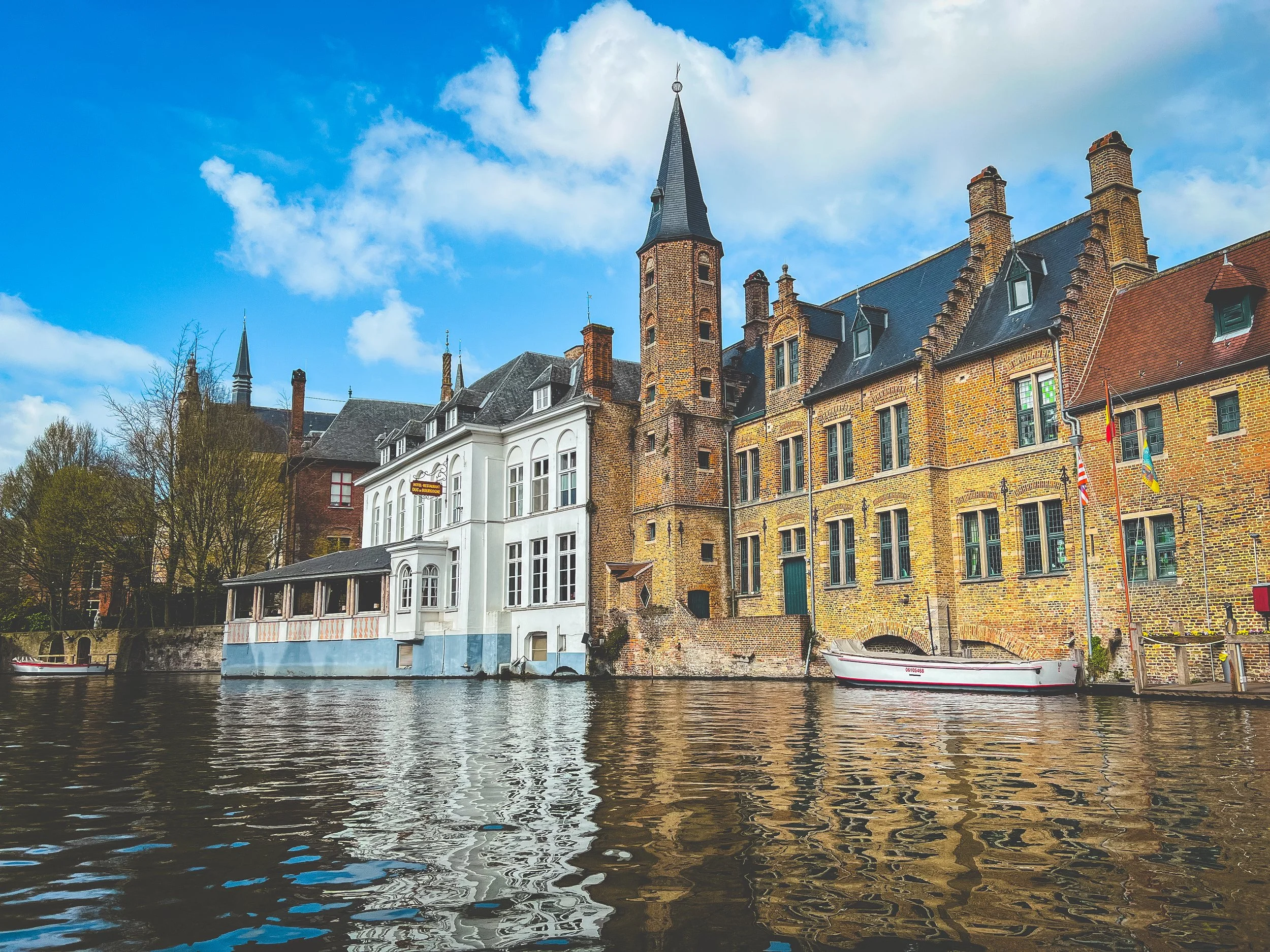 View of historic buildings in Bruges along a canal with boats, colorful sky with clouds, and trees in the background.