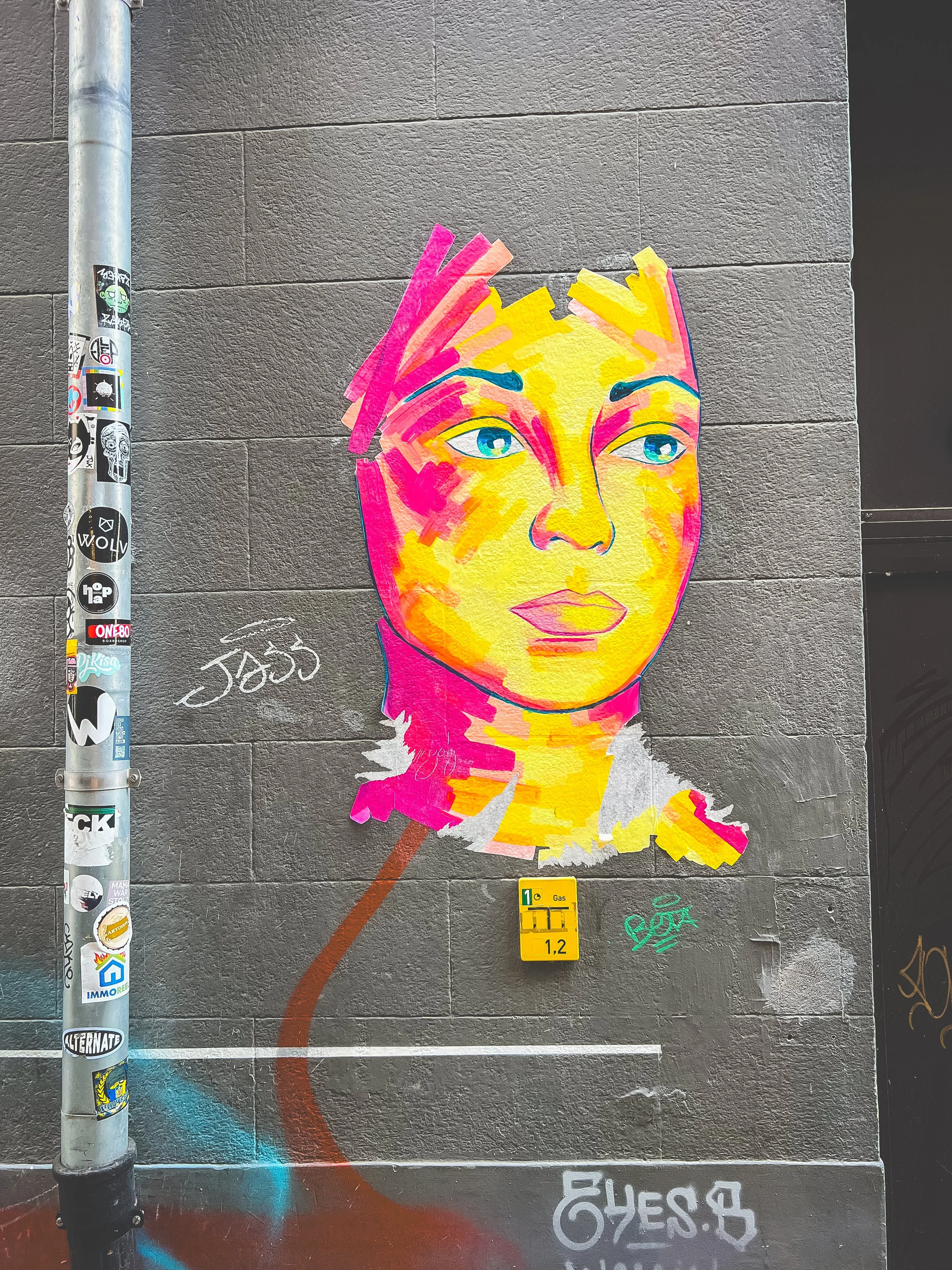 Colorful street art depicting a stylized portrait of a woman with blue eyes, yellow and pink skin tones, and pink and yellow hair on a gray wall.