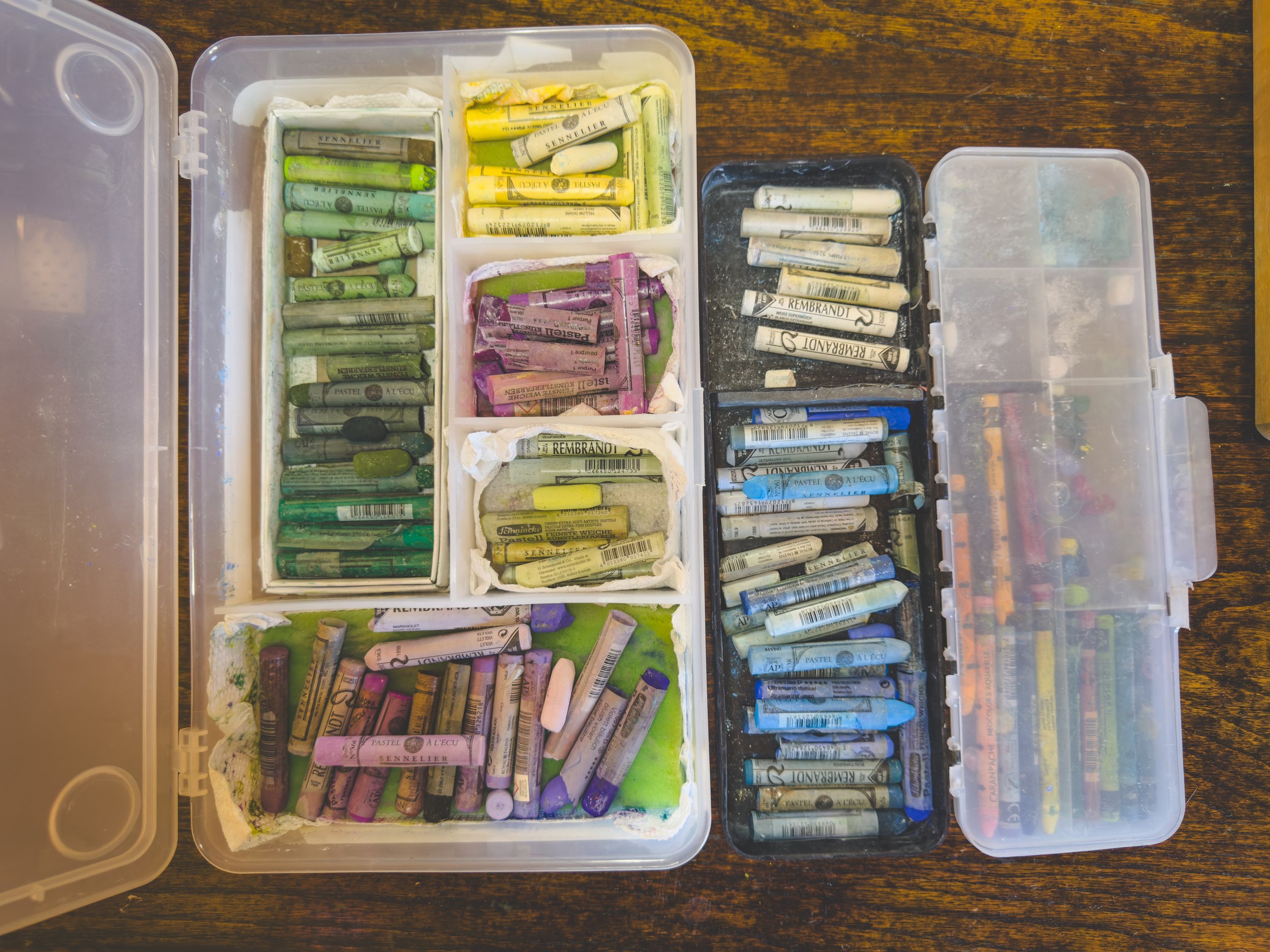 Open plastic case with multiple compartments containing various used pastel chalks organized by color, including greens, yellows, purples, blues, and pinks, on a wooden surface.